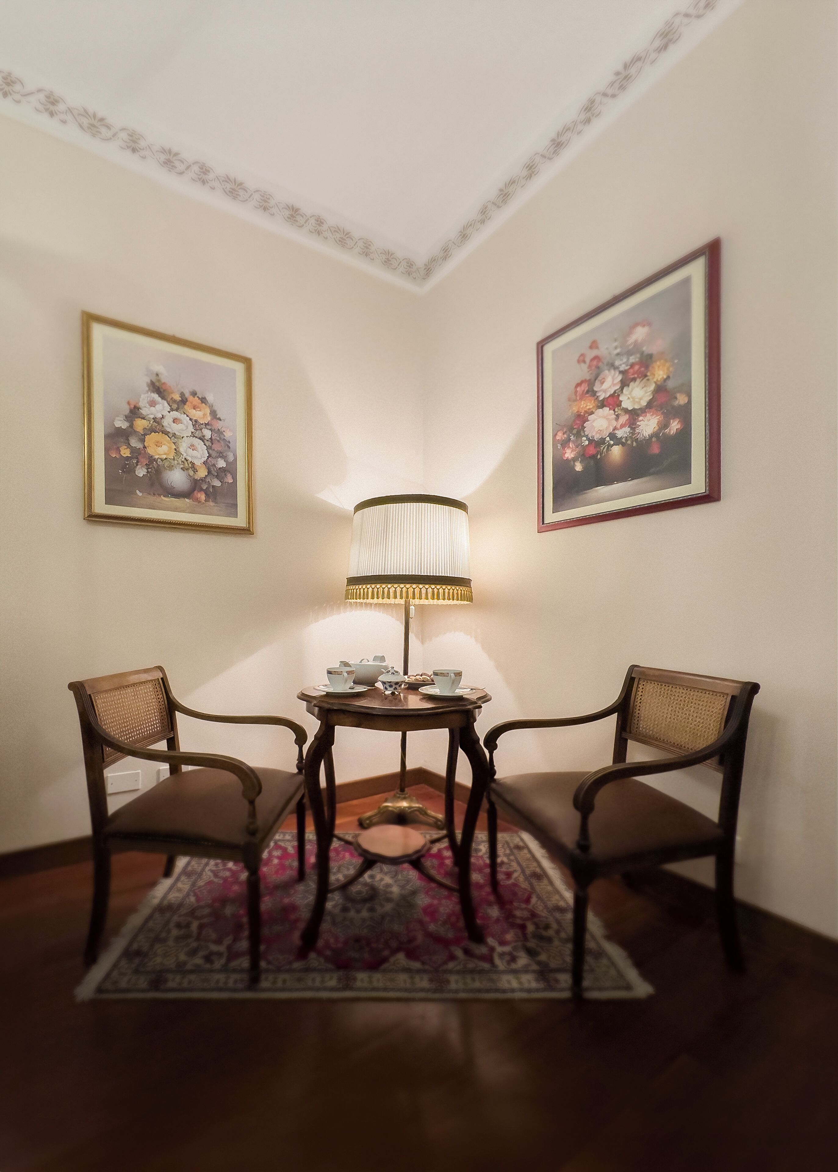 A corner room with two chairs, a small table with a lamp, and two framed floral paintings on the walls.
