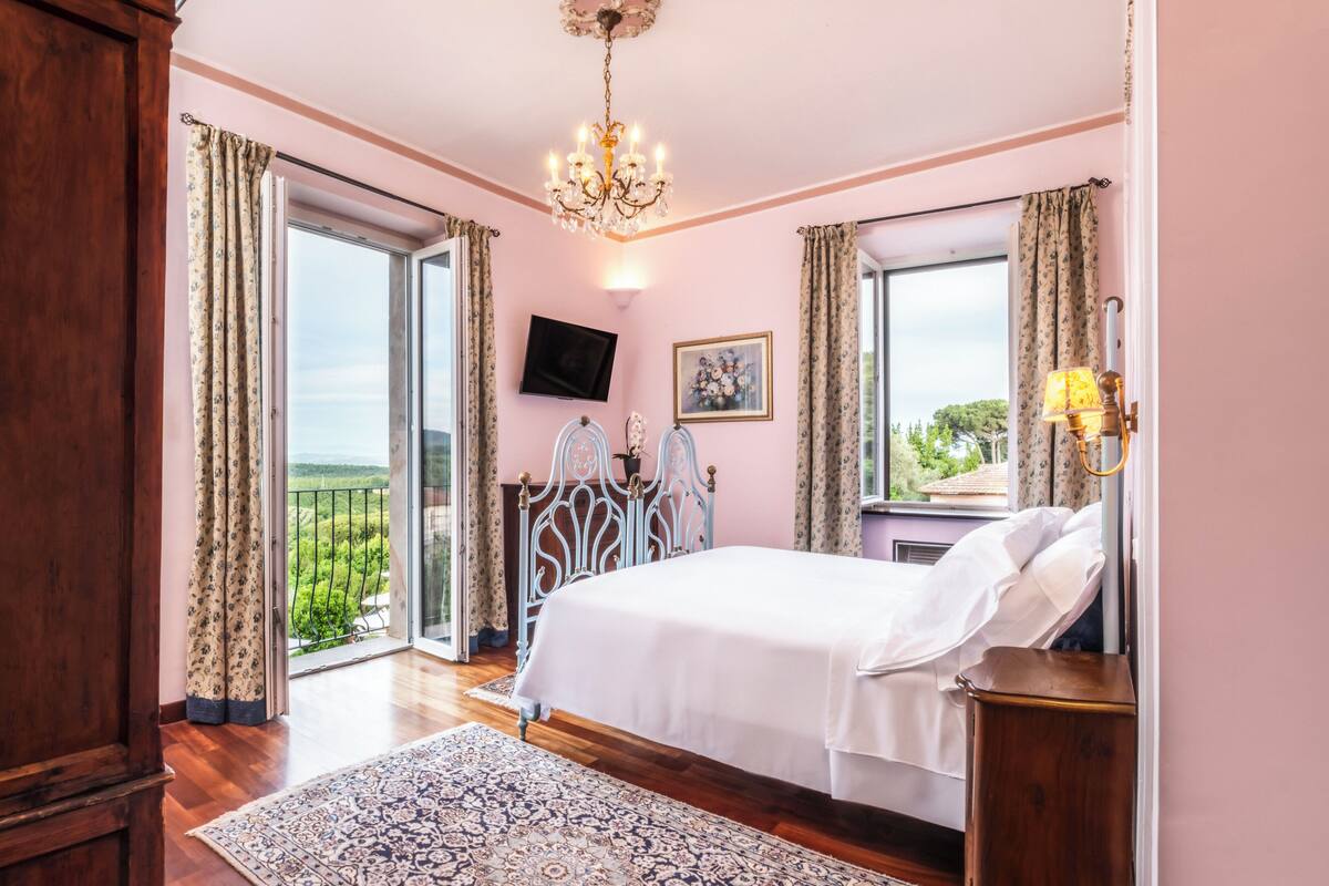 A bedroom with a bed, a chandelier, a TV, and a balcony with curtains.