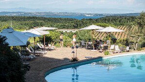 A pool area with lounge chairs and umbrellas, surrounded by greenery and a view of distant mountains. - Villa Clodia (Manziana)
