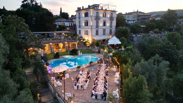 An outdoor dining area with a pool, surrounded by trees and a large building in the background. - Villa Clodia (Manziana)