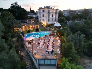 Zona pranzo all'aperto con piscina, circondata da alberi e un grande edificio sullo sfondo.