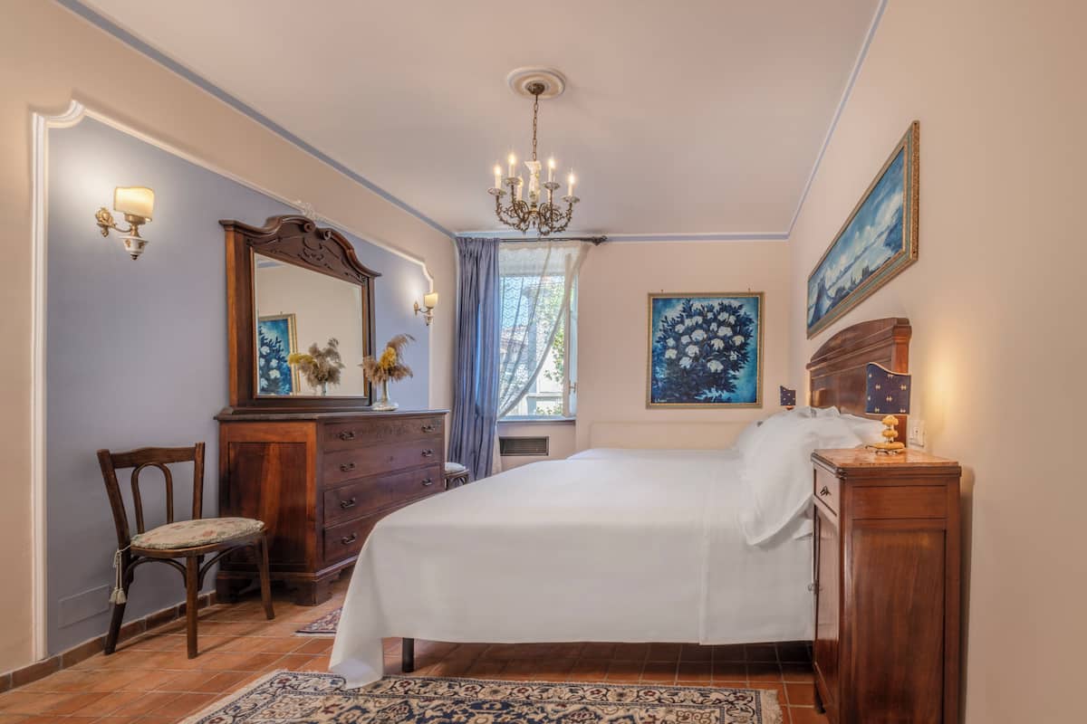 A bedroom with a large bed, a wooden dresser, a chair, a chandelier, and a painting on the wall.