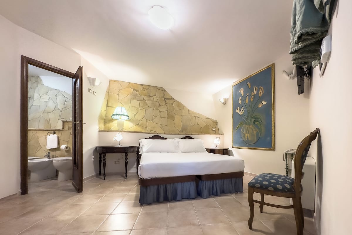 A bedroom with a bed, a chair, a painting, a bathroom with a mirror, and a stone wall.