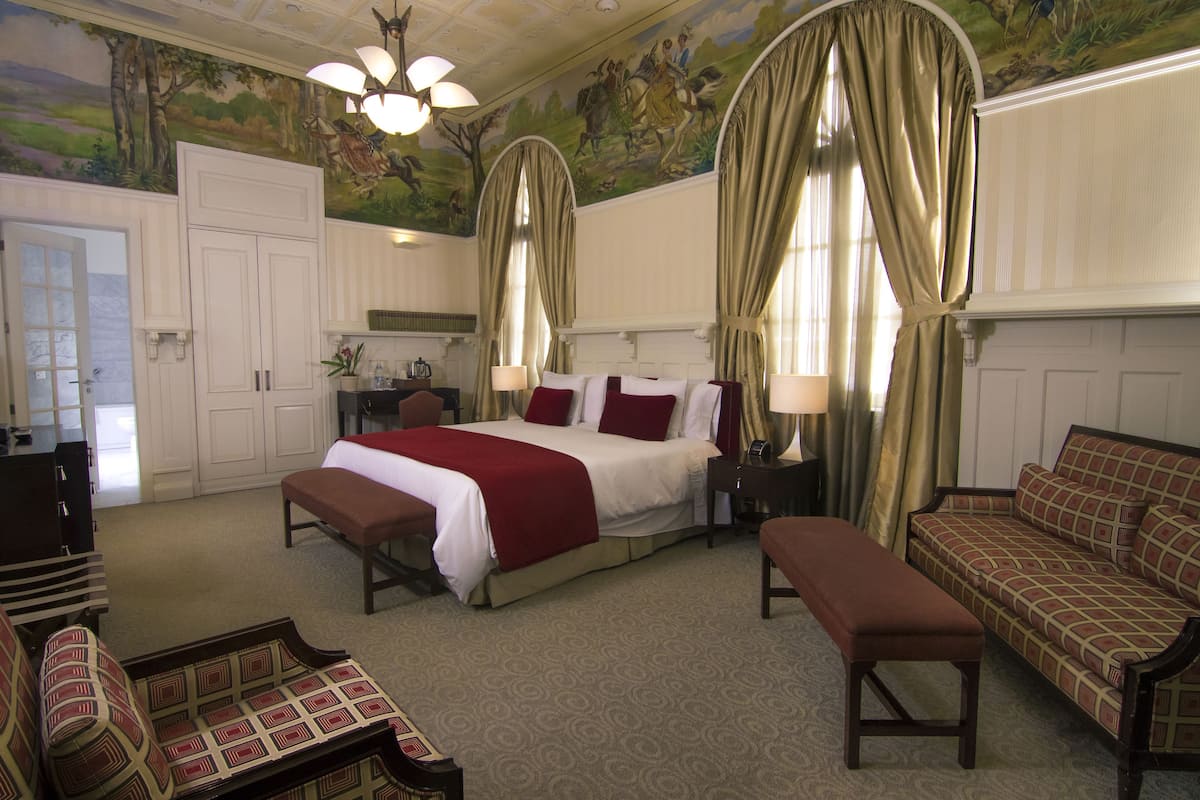 A hotel room with a large bed, a sofa, a bench, and a desk with a lamp.