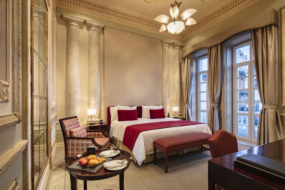 A hotel room with a large bed, a dining table with fruits, a bench, and a view of the city through large windows.
