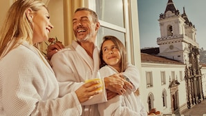 A couple and a child in a white robe, holding a glass of orange juice, with a historic building in the background. - Casa Gangotena (Quito)