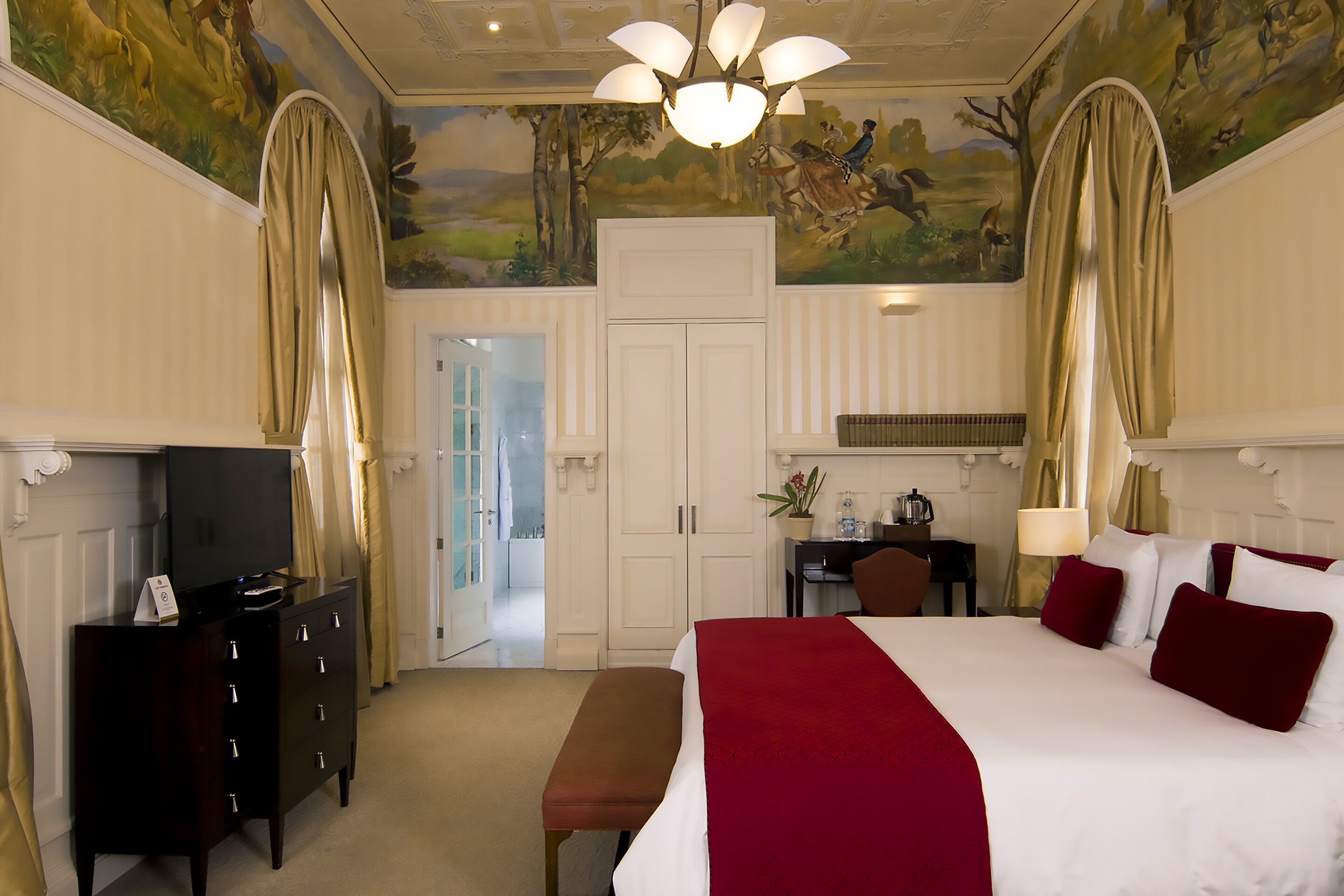 Habitación de hotel con una cama grande, un televisor en una cómoda, un escritorio con una lámpara y un mural en el techo.
