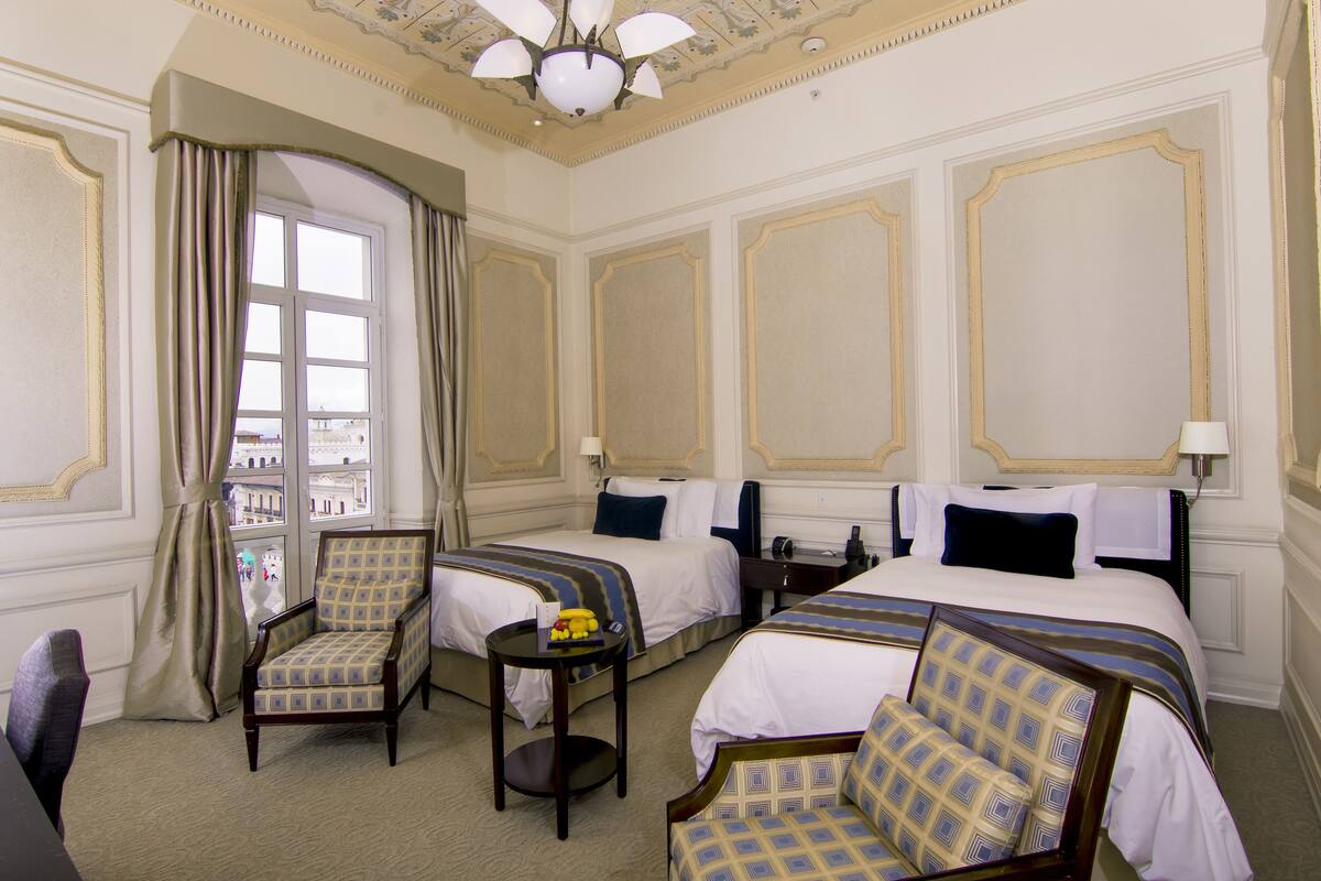 A hotel room with two beds, a sitting area with a chair and a small table, and a view through a window.