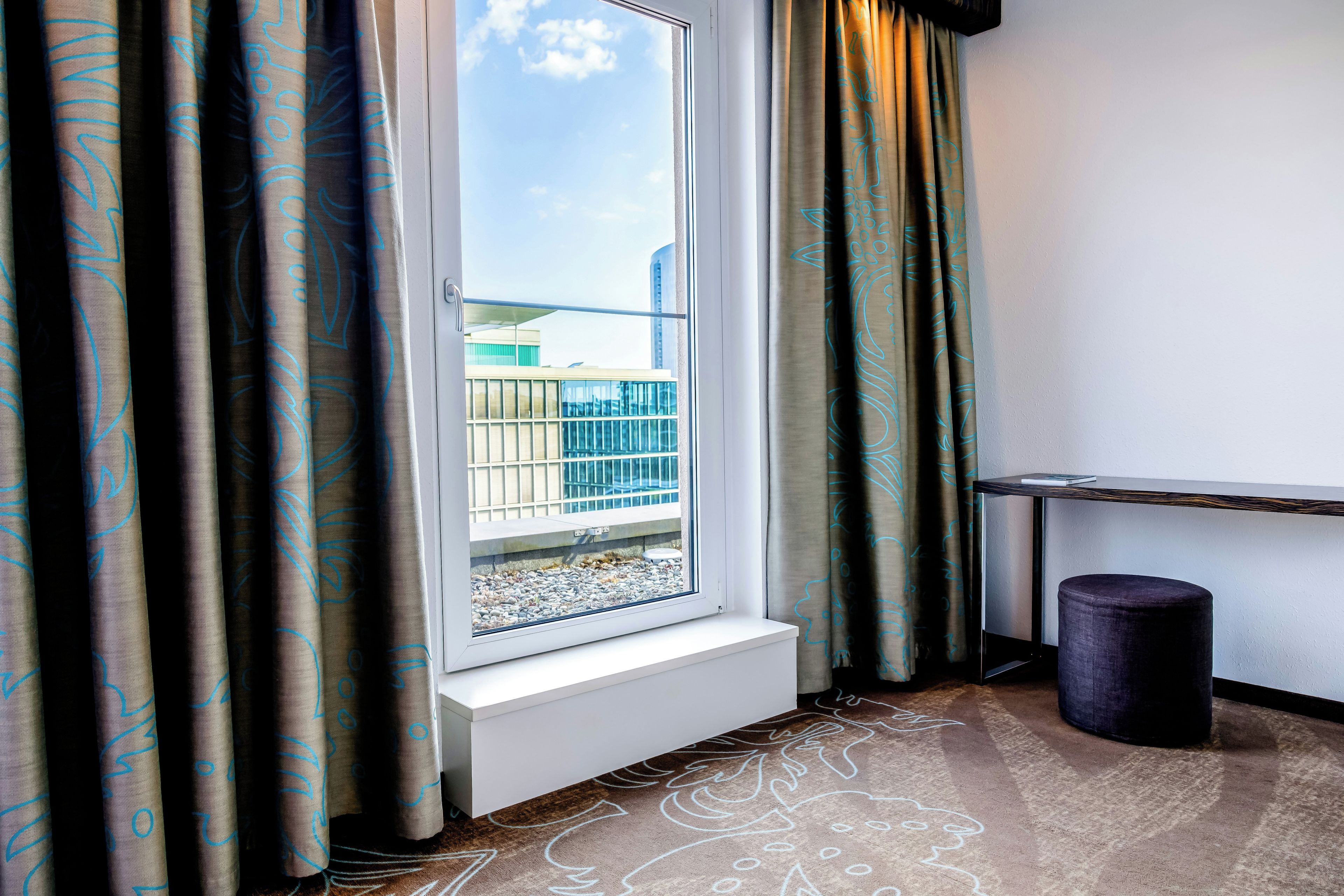 Une chambre d’hôtel avec une fenêtre, des rideaux, une petite table et une poubelle.