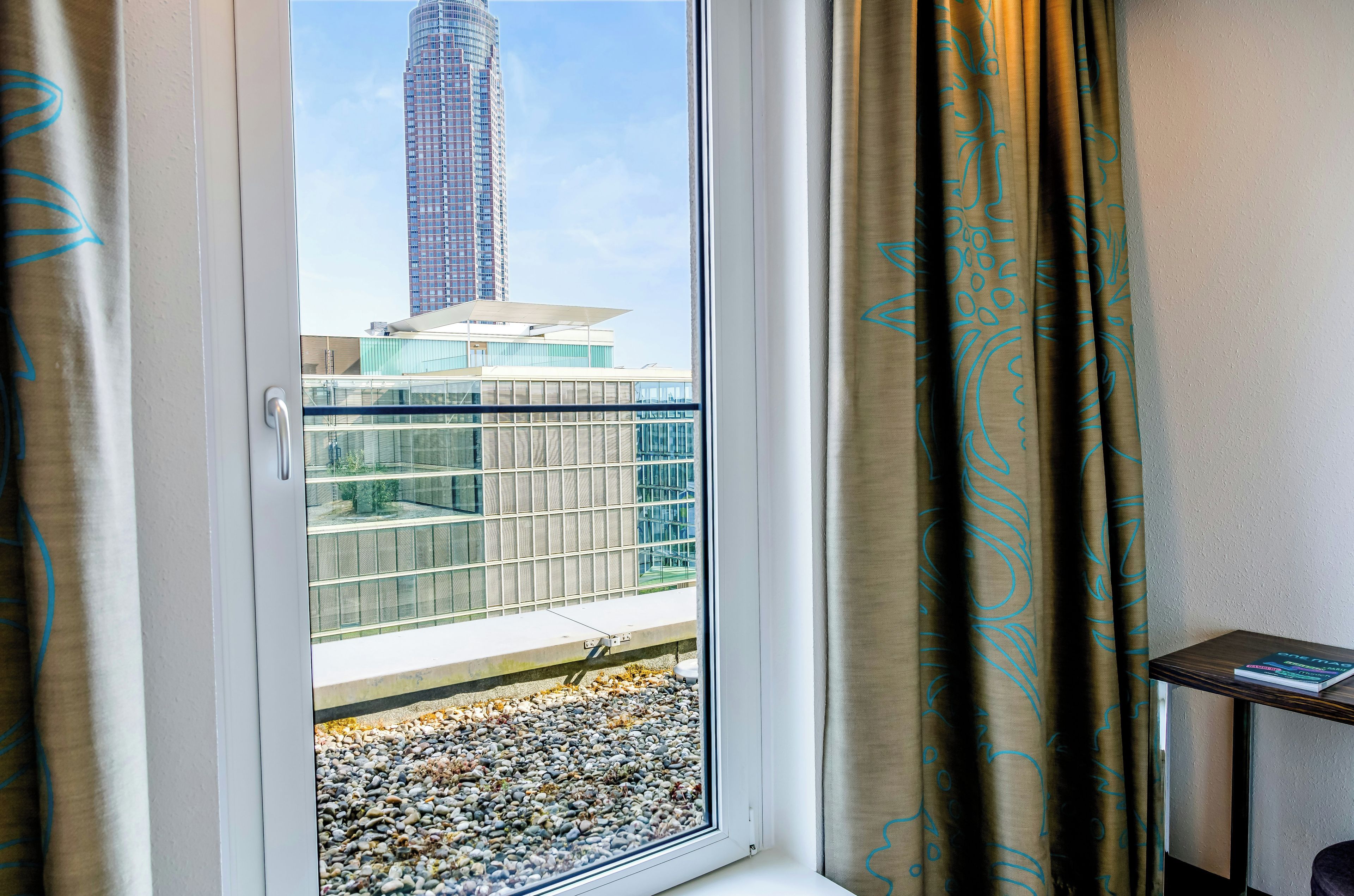 Ein Fenster mit Blick auf ein hohes Gebäude, ein Balkon mit Glastrennwand und ein kleiner Tisch mit einem Buch.