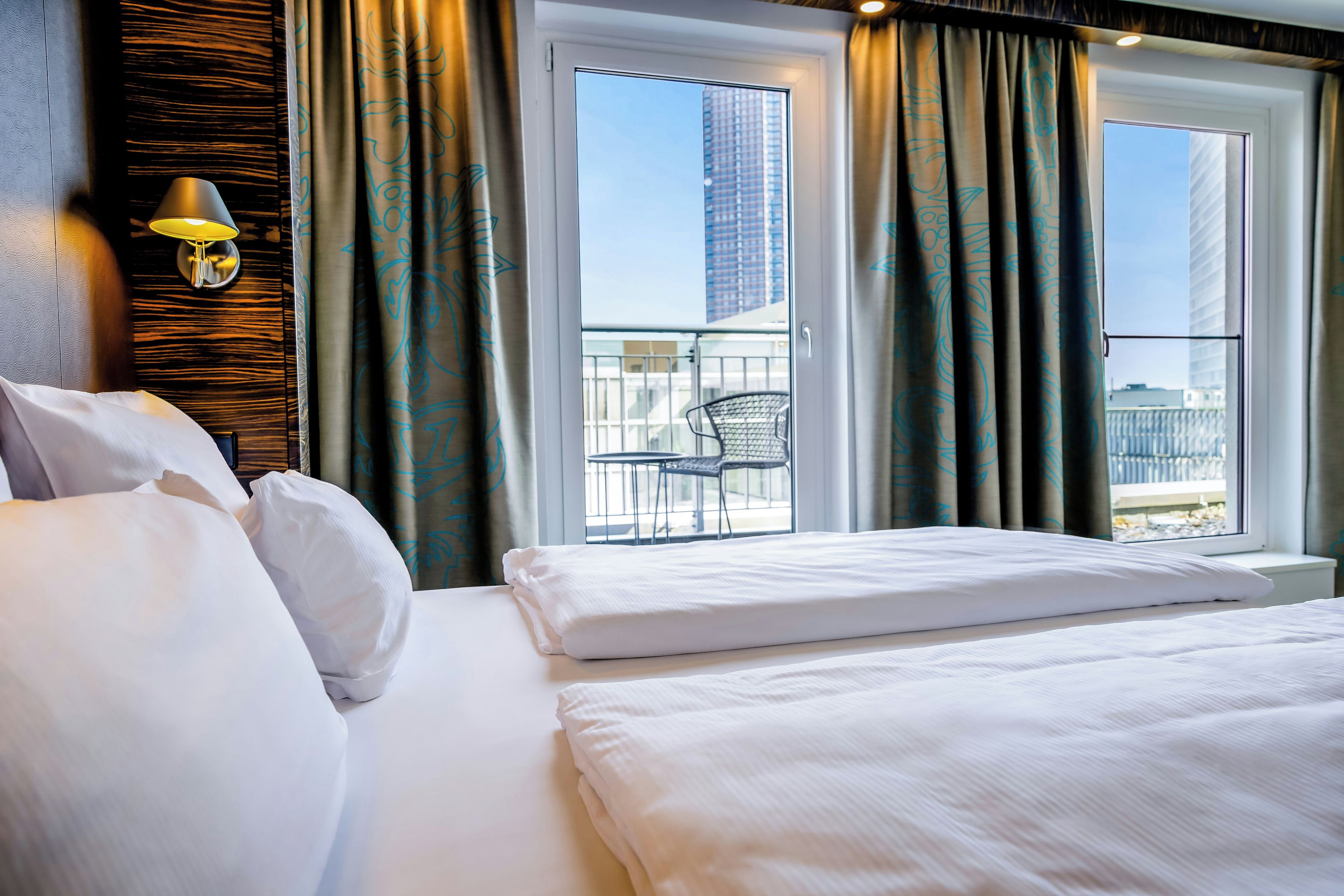 A hotel room with a bed, a window with curtains, and a view of a balcony with a chair.