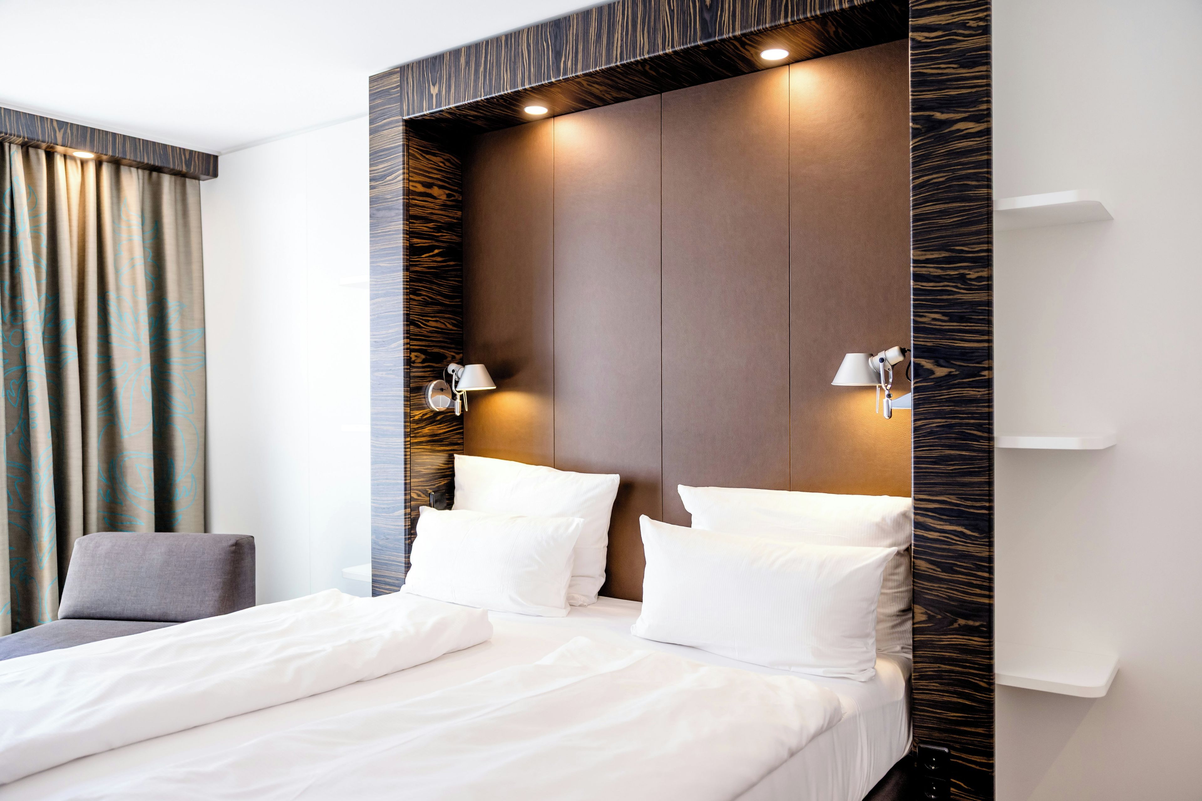 Une chambre d’hôtel moderne avec un grand lit, deux lampes de chevet et une tête de lit à motifs évoquant le bois.
