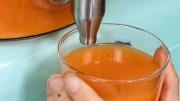 A hand holding a glass with a clear liquid, possibly tea, being poured from a metallic container.