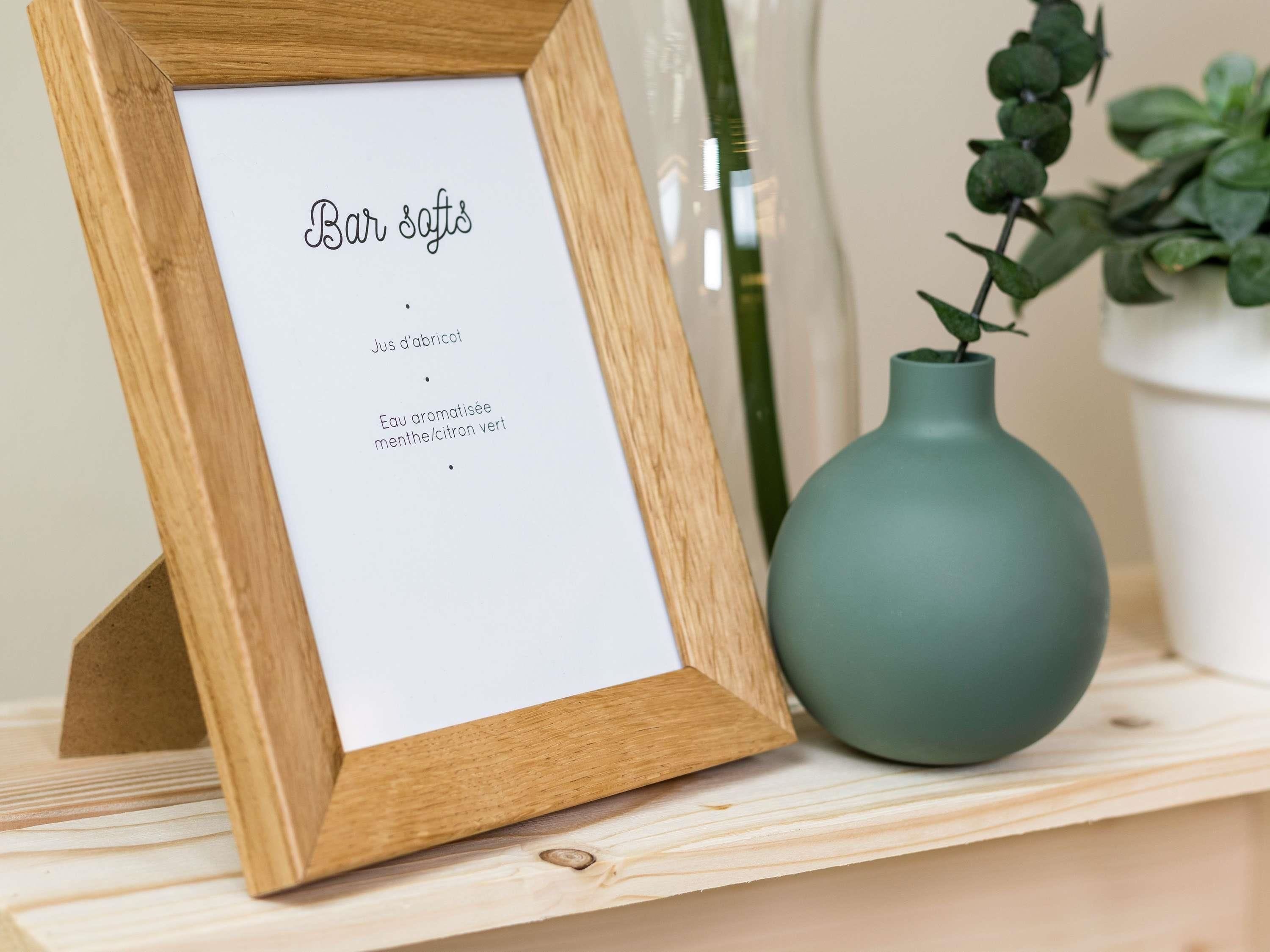 A wooden-framed sign with text in a foreign language, a teal vase with a plant, and a white pot in the background.