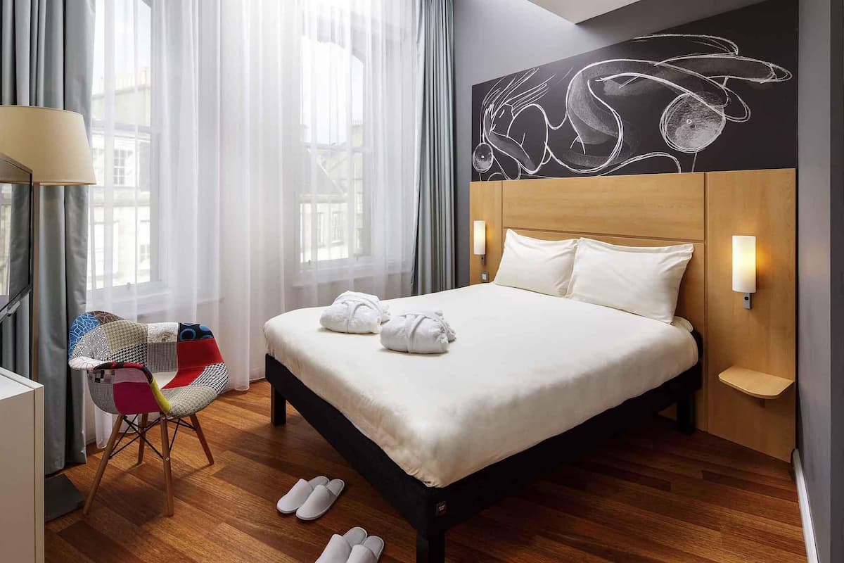 Ein Hotelzimmer mit Bett, einem Sessel, einem Wandbild und einem Fenster mit Vorhängen.