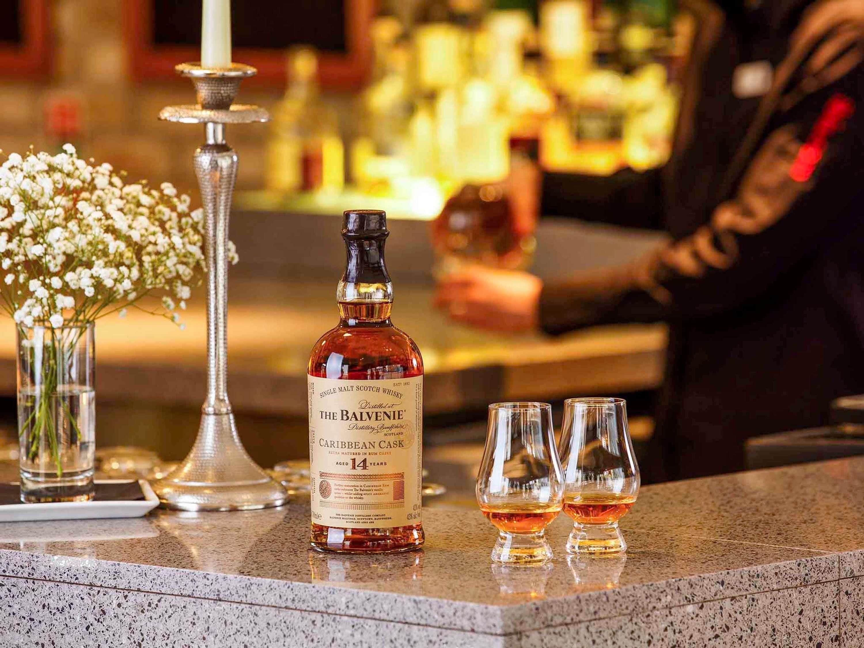 A bottle of The Balvenie Caribbean Cask whisky, two glasses, and a candle on a bar counter.