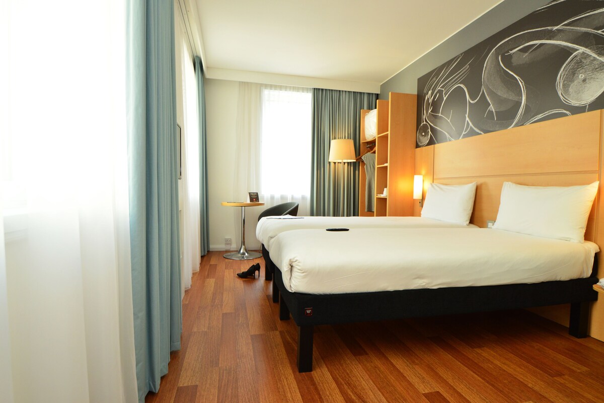 Habitación de hotel con una cama grande, un escritorio, una lámpara y vistas a la ciudad desde la ventana.