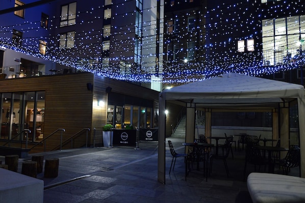 An outdoor seating area with tables and chairs, a covered section, and string lights overhead.