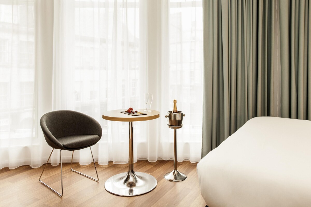 Une chambre d’hôtel avec un lit, une table ronde sur laquelle se trouve une bouteille de champagne, une chaise et des rideaux légers.