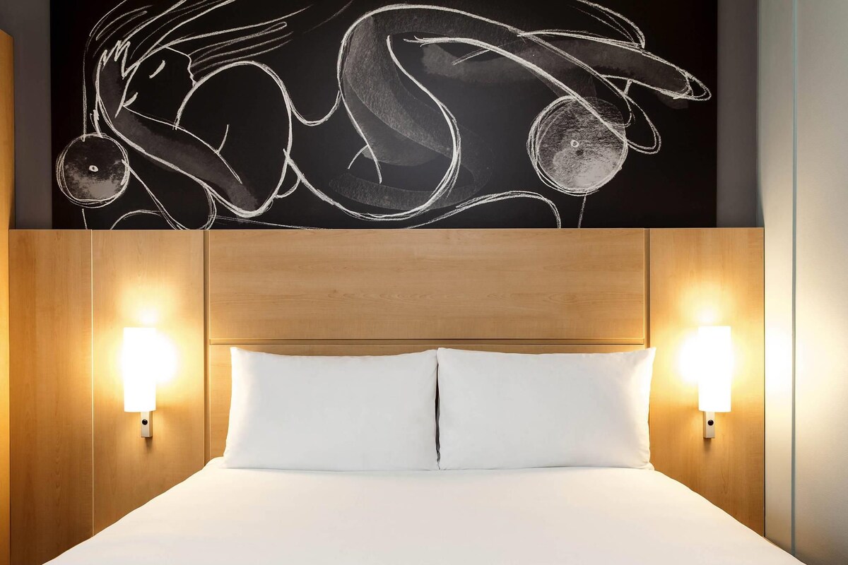 Une chambre d’hôtel avec une tête de lit en bois, deux lampes de chevet et une grande œuvre d’art abstraite au-dessus du lit.
