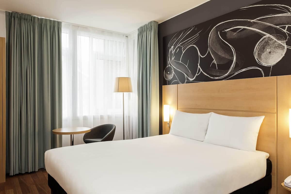 Ein Hotelzimmer mit einem großen Bett, einem Schreibtisch, einem Sessel und einer Wand mit abstrakter Kunst.