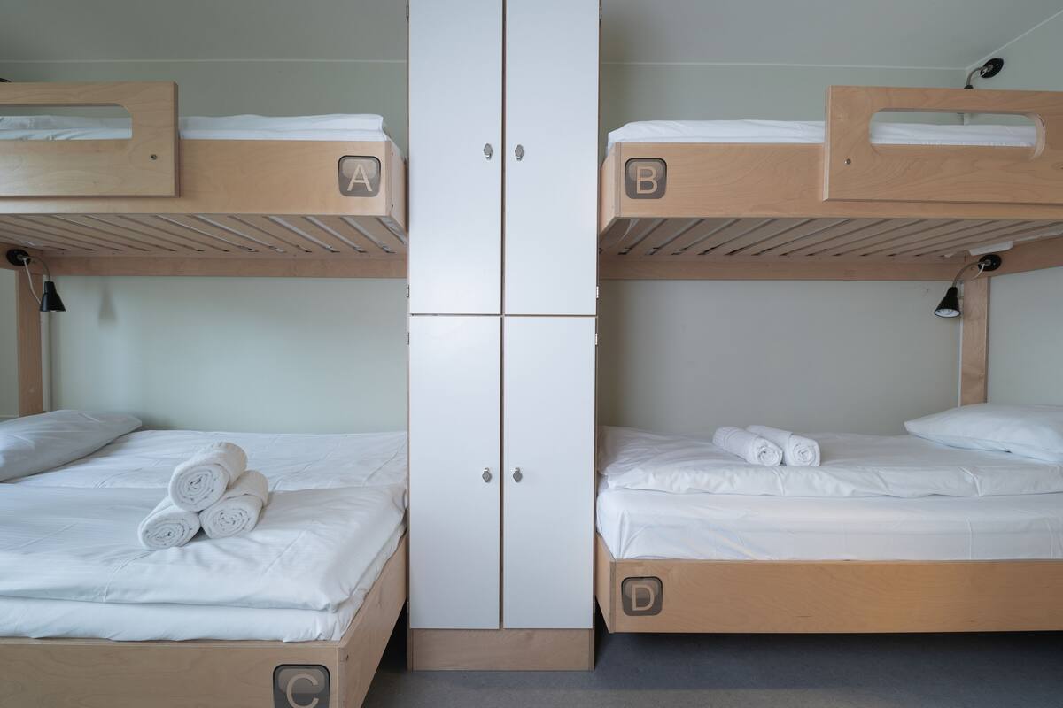 A room with two bunk beds, each with a wooden headboard and a white mattress. There are two sets of drawers below the beds, and a white wardrobe in the center.