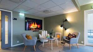 A modern lounge area with a flat-screen TV displaying a fire, two armchairs, a small table, and a floor lamp. - Rumi Hostel (Trondheim)