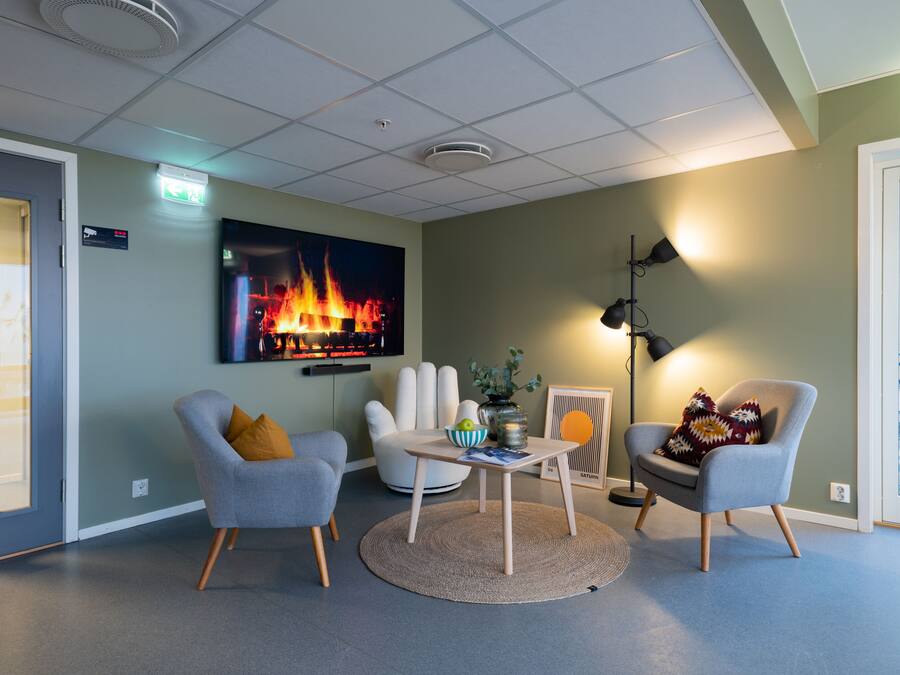 Un área de estar moderna con una televisión de pantalla plana mostrando un fuego, dos sillones, una mesita y una lámpara de pie.