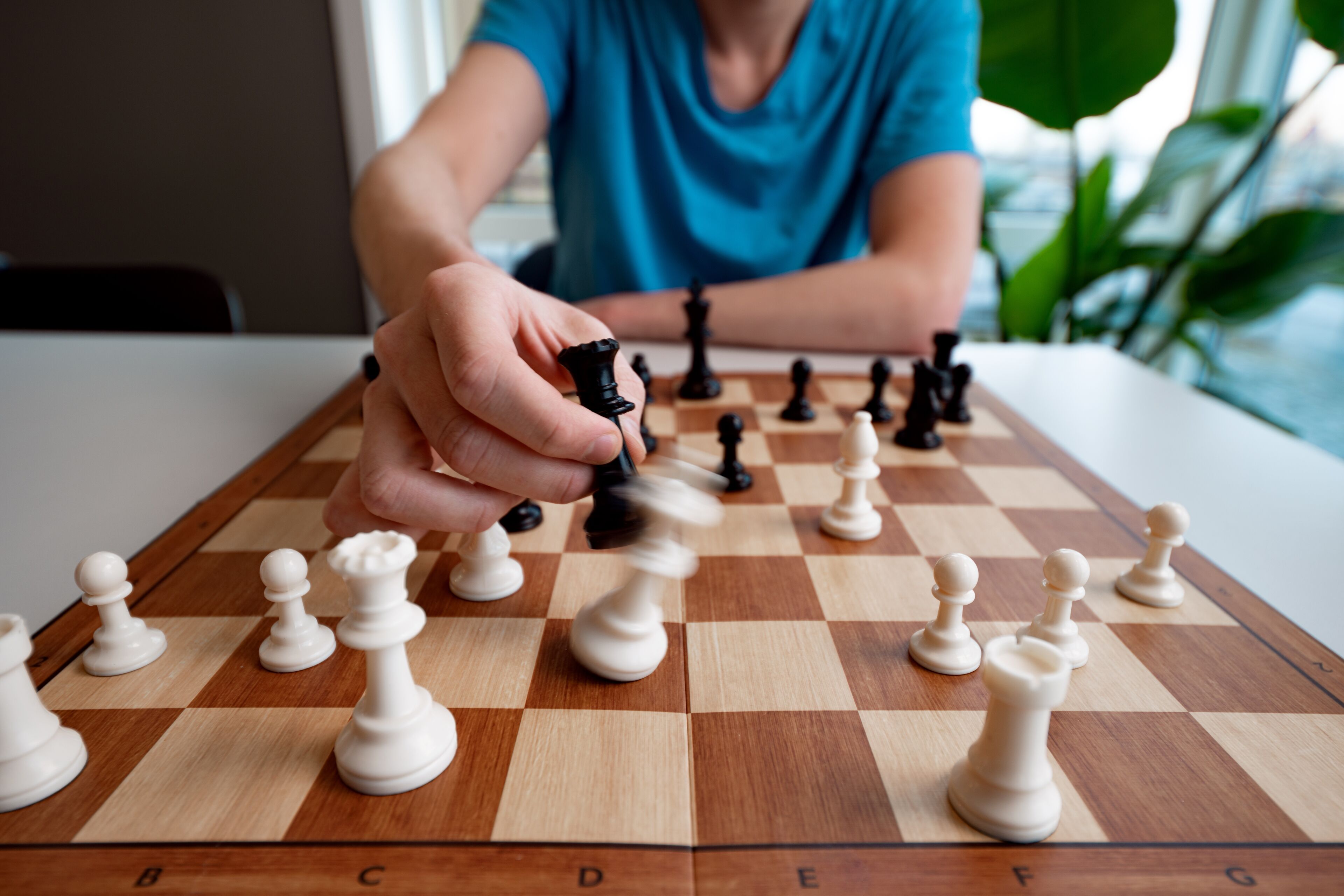 A chessboard with pieces in a game, a hand moving a piece, and a person in a blue shirt in the background.