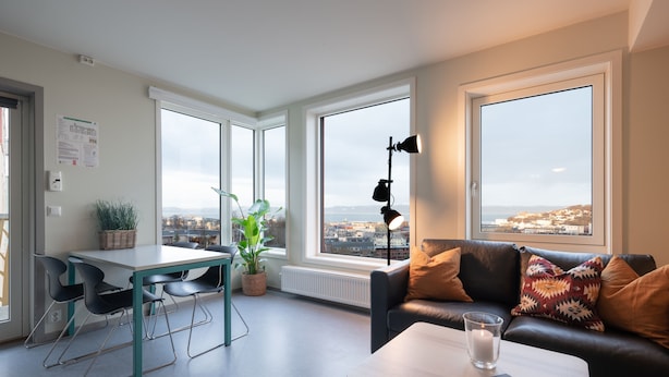 A modern living room with a dining area, large windows with a city view, a black leather sofa, and a table with a glass of water. - Rumi Hostel (Trondheim)