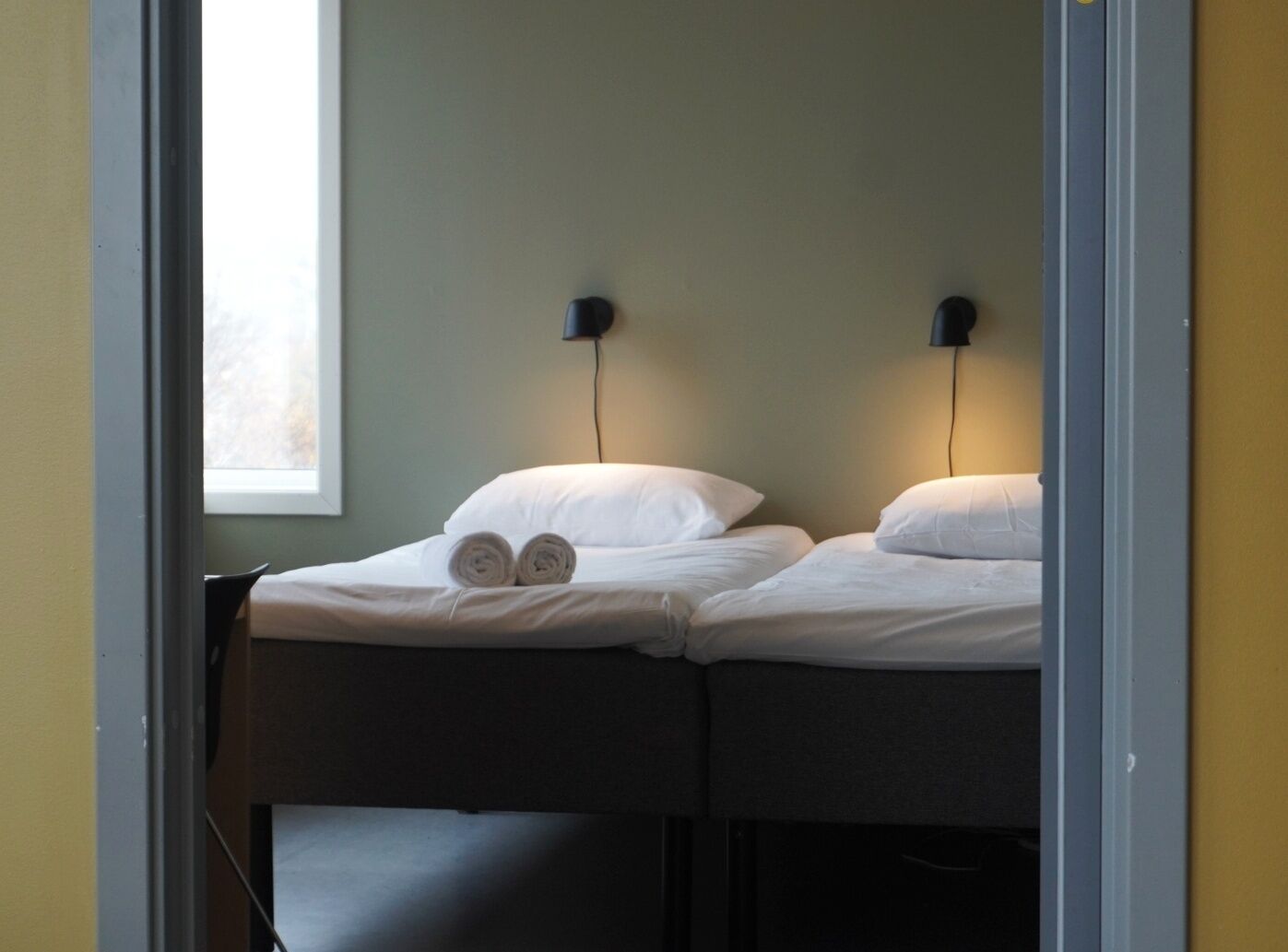 A room with two single beds, each with a pillow and a rolled towel. There are two wall-mounted nightstands with lamps.