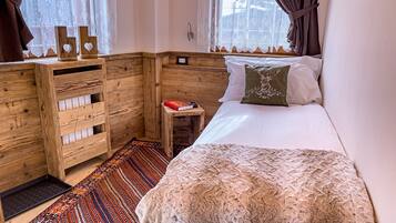 A bedroom with a bed, a small bedside table, a wooden wardrobe, patterned rugs, and a window with curtains.