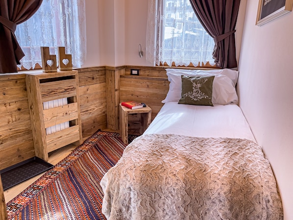 A bedroom with a bed, a small bedside table, a wooden wardrobe, patterned rugs, and a window with curtains. - Hotel da Beppe Sello (Cortina d'Ampezzo)