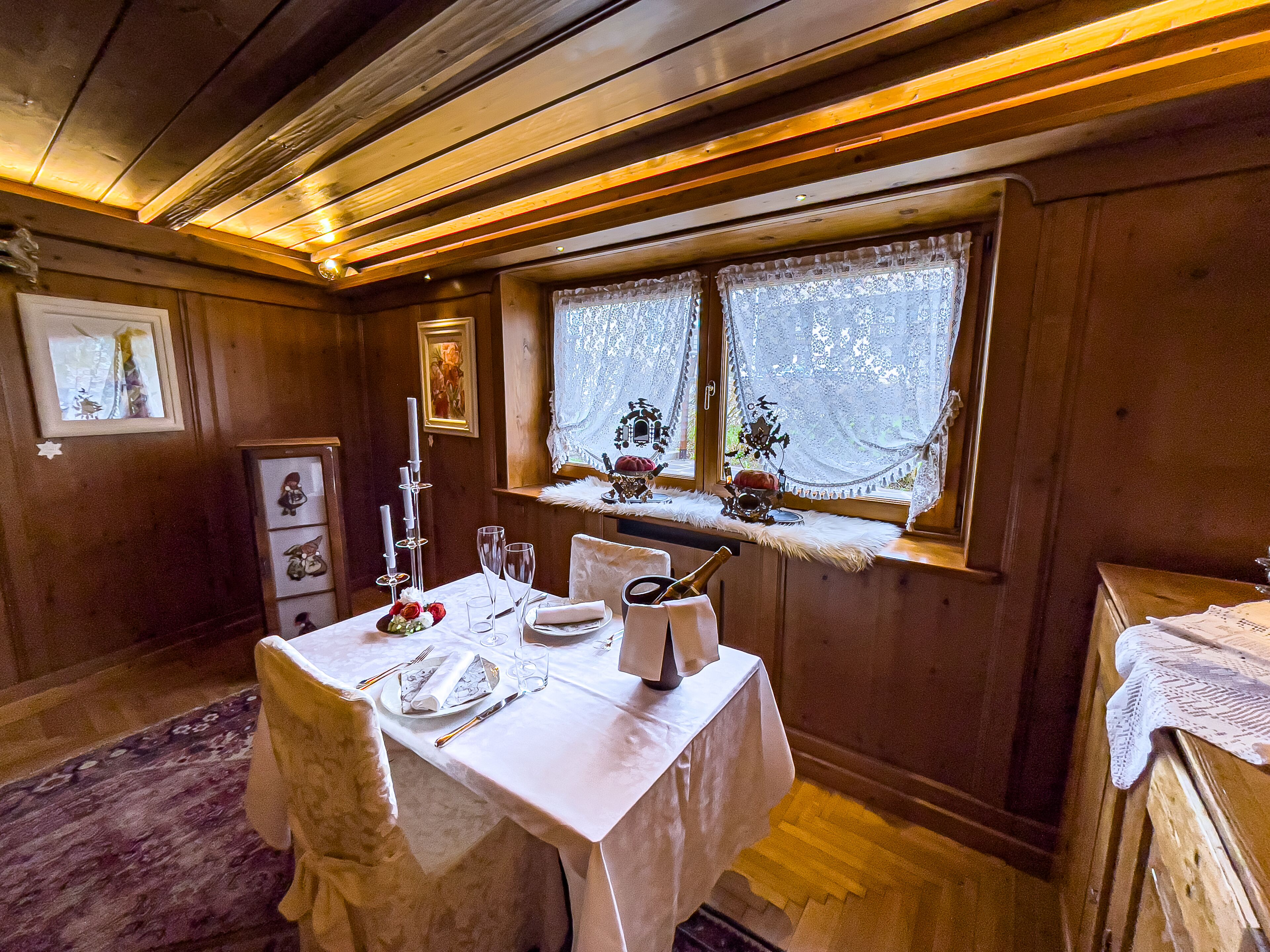 A dining area with a wooden interior, a table set for two, and a window with lace curtains.