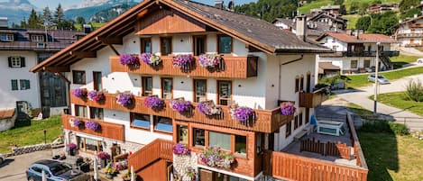 Un immeuble à plusieurs étages, doté de balcons ornés de jardinières fleuries, d’un patio avec des sièges extérieurs et offrant une vue sur un paysage montagneux en arrière-plan.