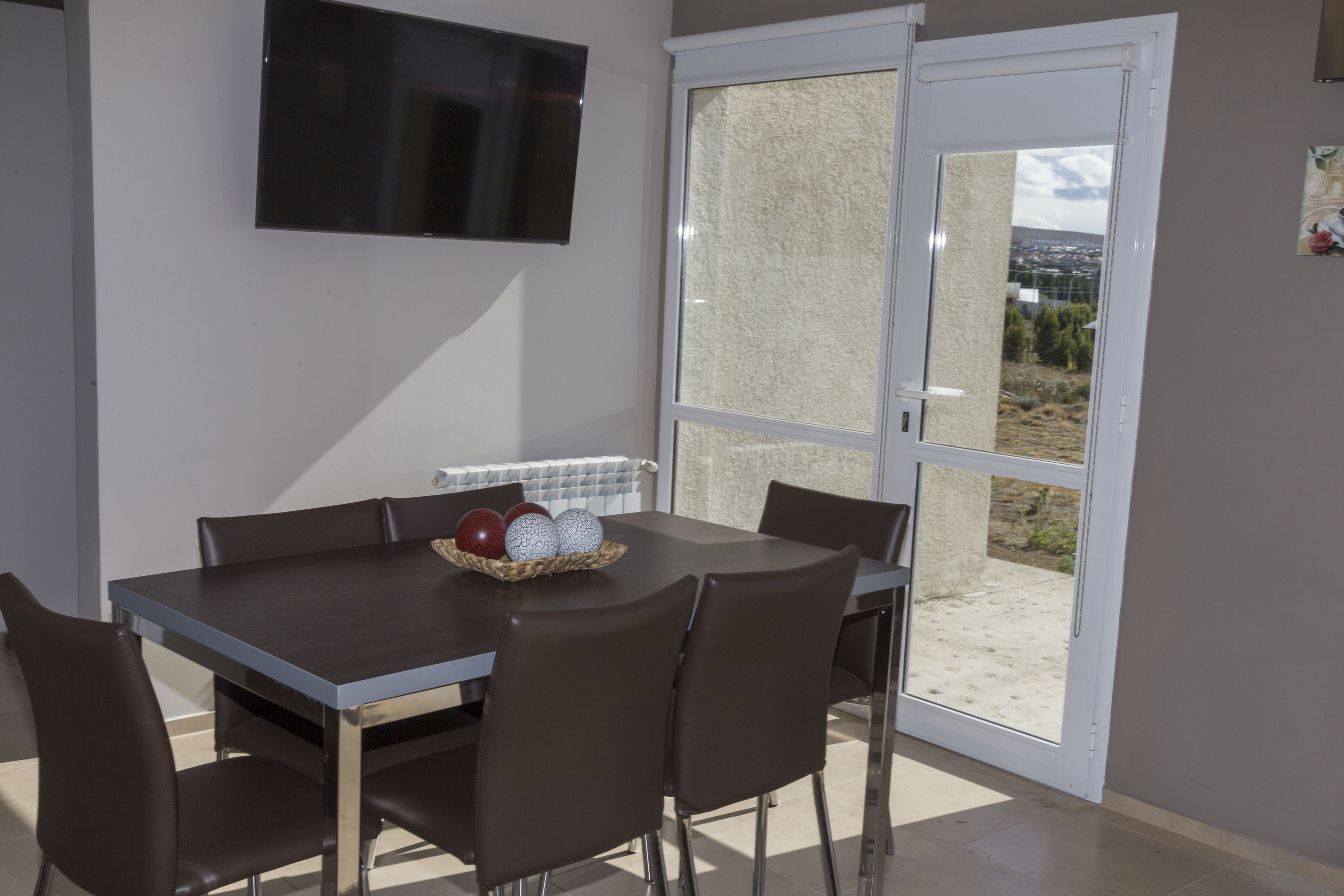 A dining area with a table and chairs, a television mounted on the wall, and a sliding glass door leading to an outdoor area.