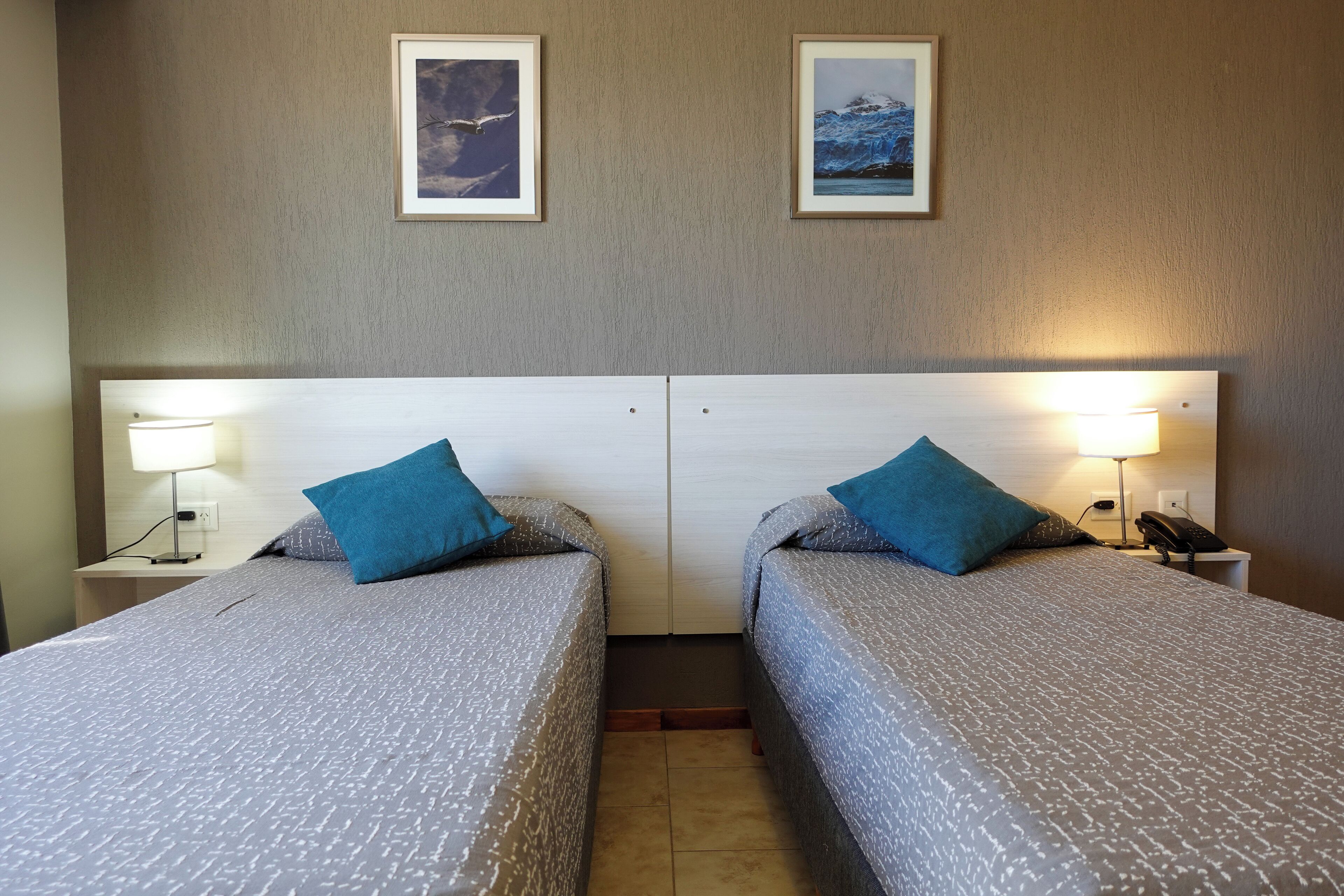 Ein Hotelzimmer mit zwei Betten, jedes mit einem blauen Kissen, einem Nachttisch mit Lampe sowie gerahmten Bildern an der Wand.