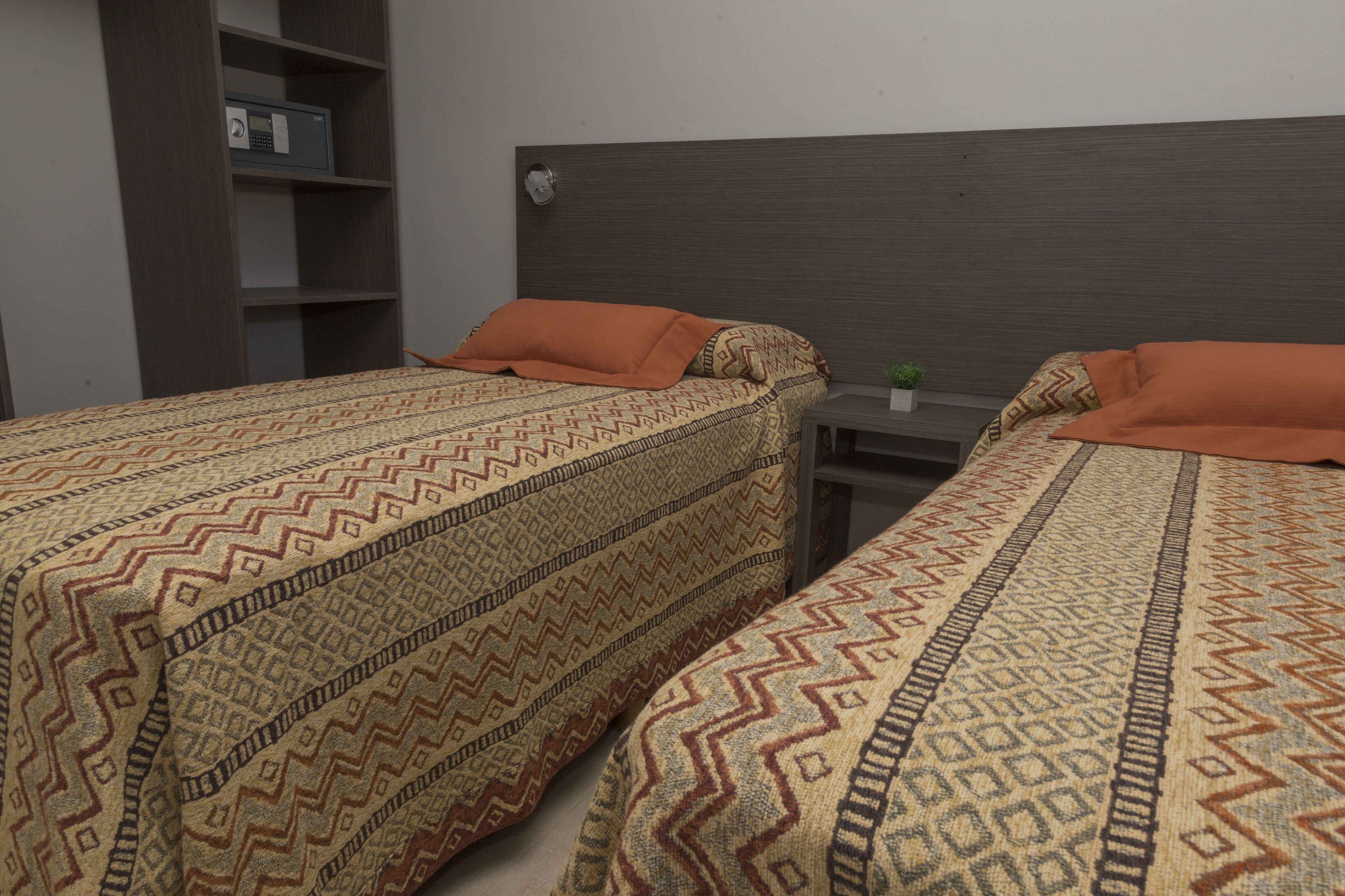 Ein Zimmer mit zwei Betten, jeweils mit gemusterten Bettbezügen und orangefarbenen Kissen. Ein kleiner Tisch mit einer Pflanze darauf und ein Regal an der Wand.