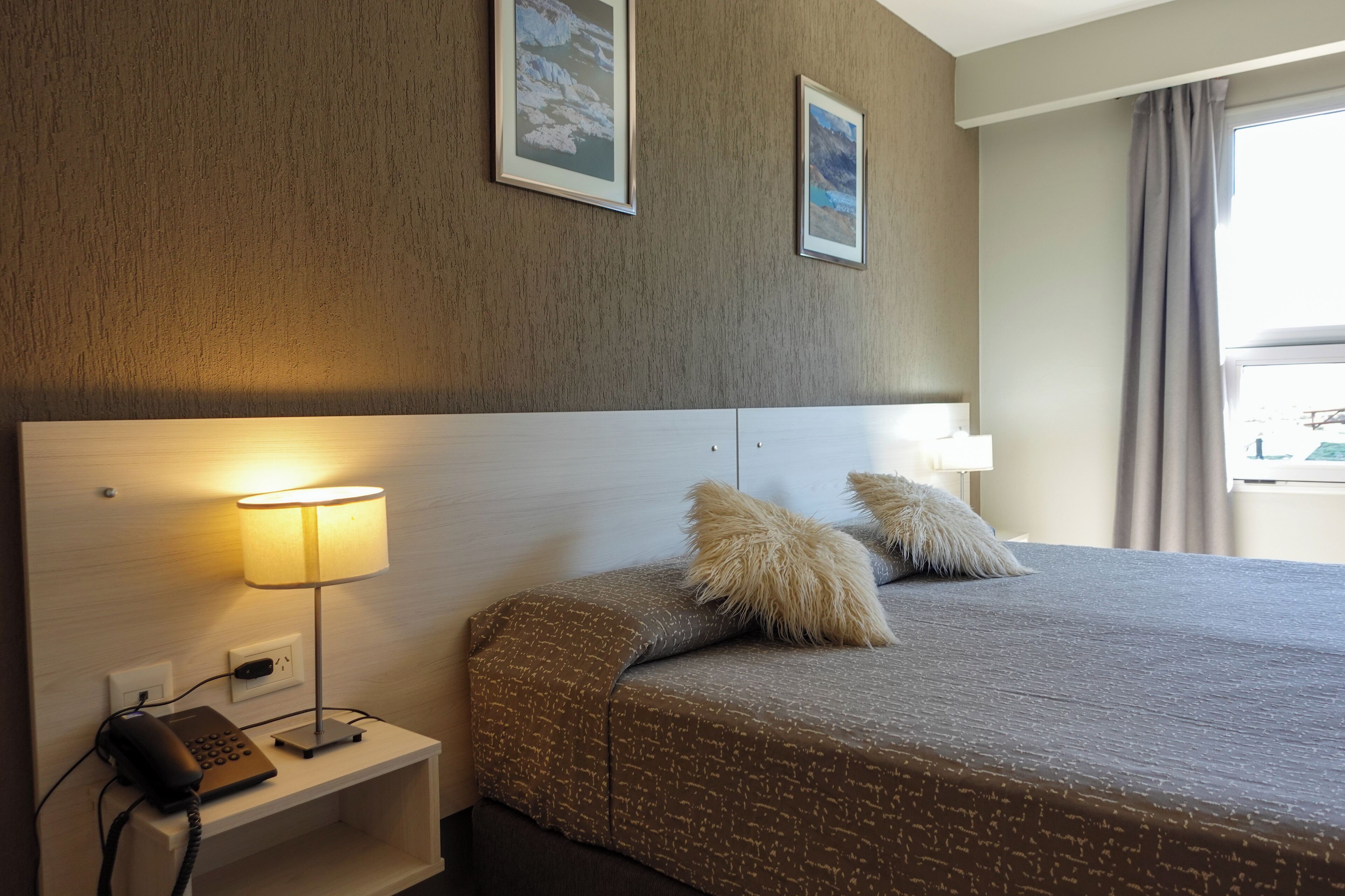 Habitación de hotel con una cama, una lámpara de noche, un teléfono y dos cuadros en la pared.