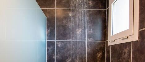 A shower with a rain showerhead, dark tiled walls, and a window.