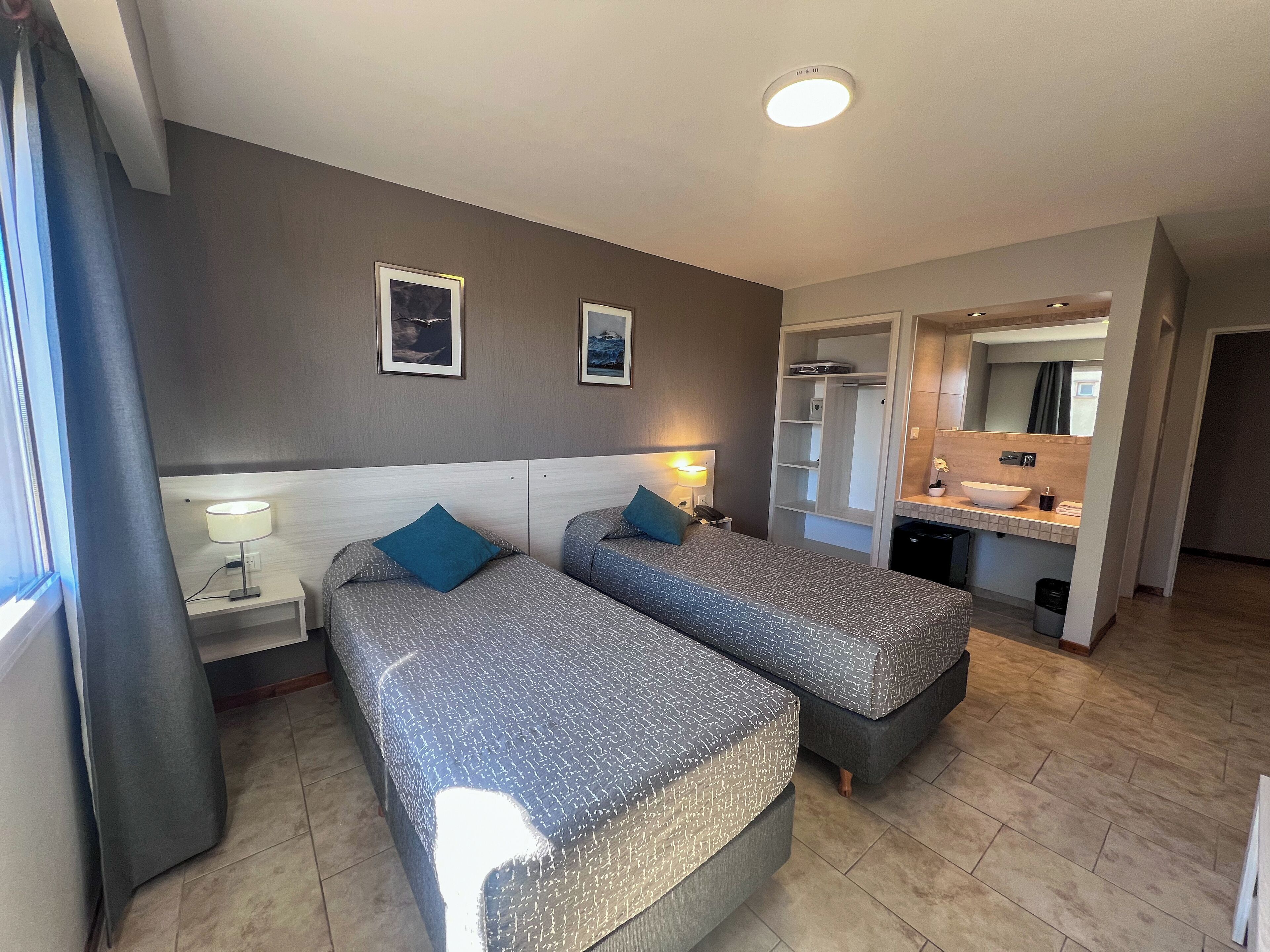 A hotel room with two beds, a nightstand with a lamp, a wardrobe, a bathroom with a sink and mirror, and a ceiling light.