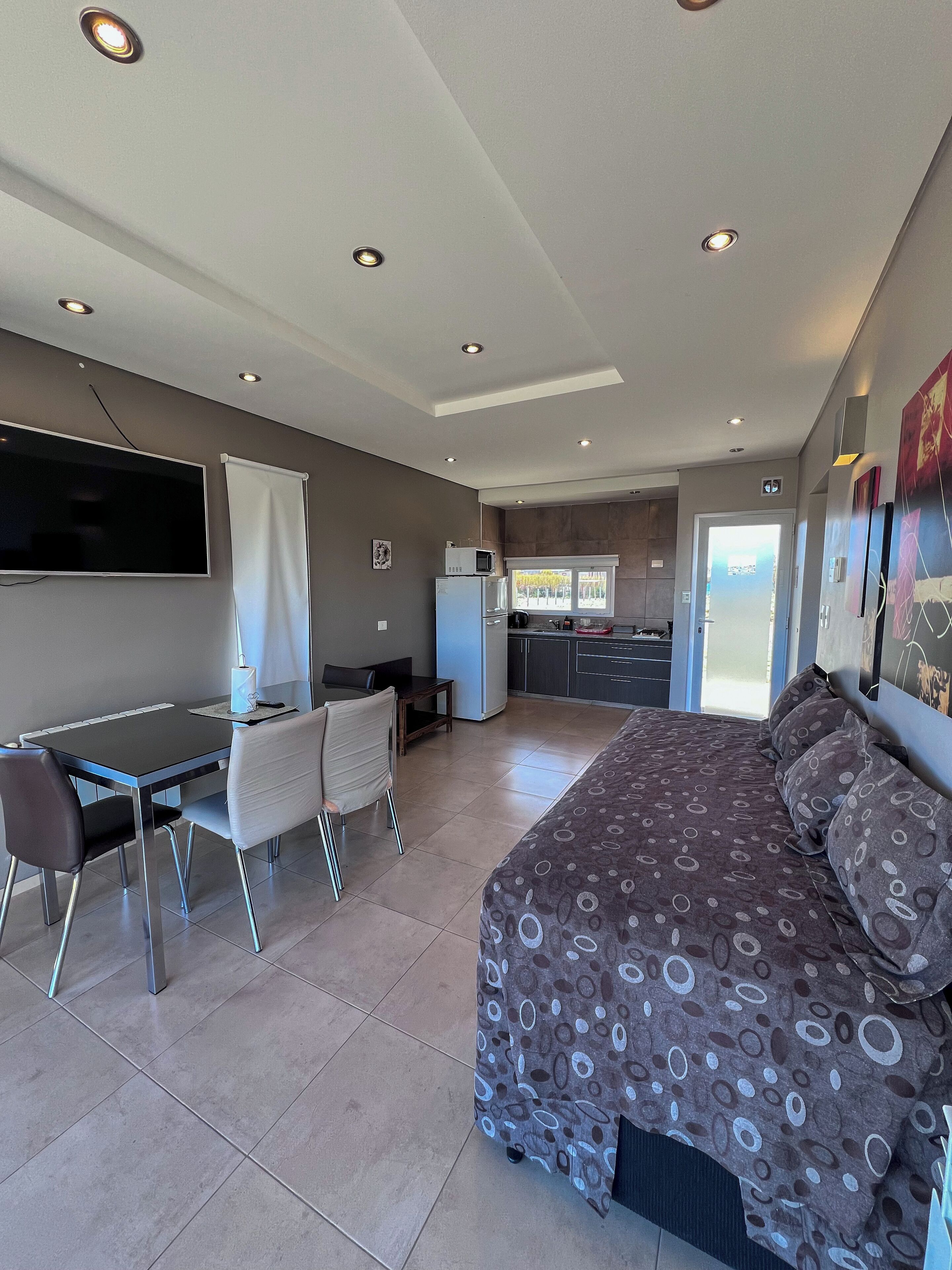 Una habitación de hotel moderna con una cama grande, un escritorio con silla, televisor y kitchenette.