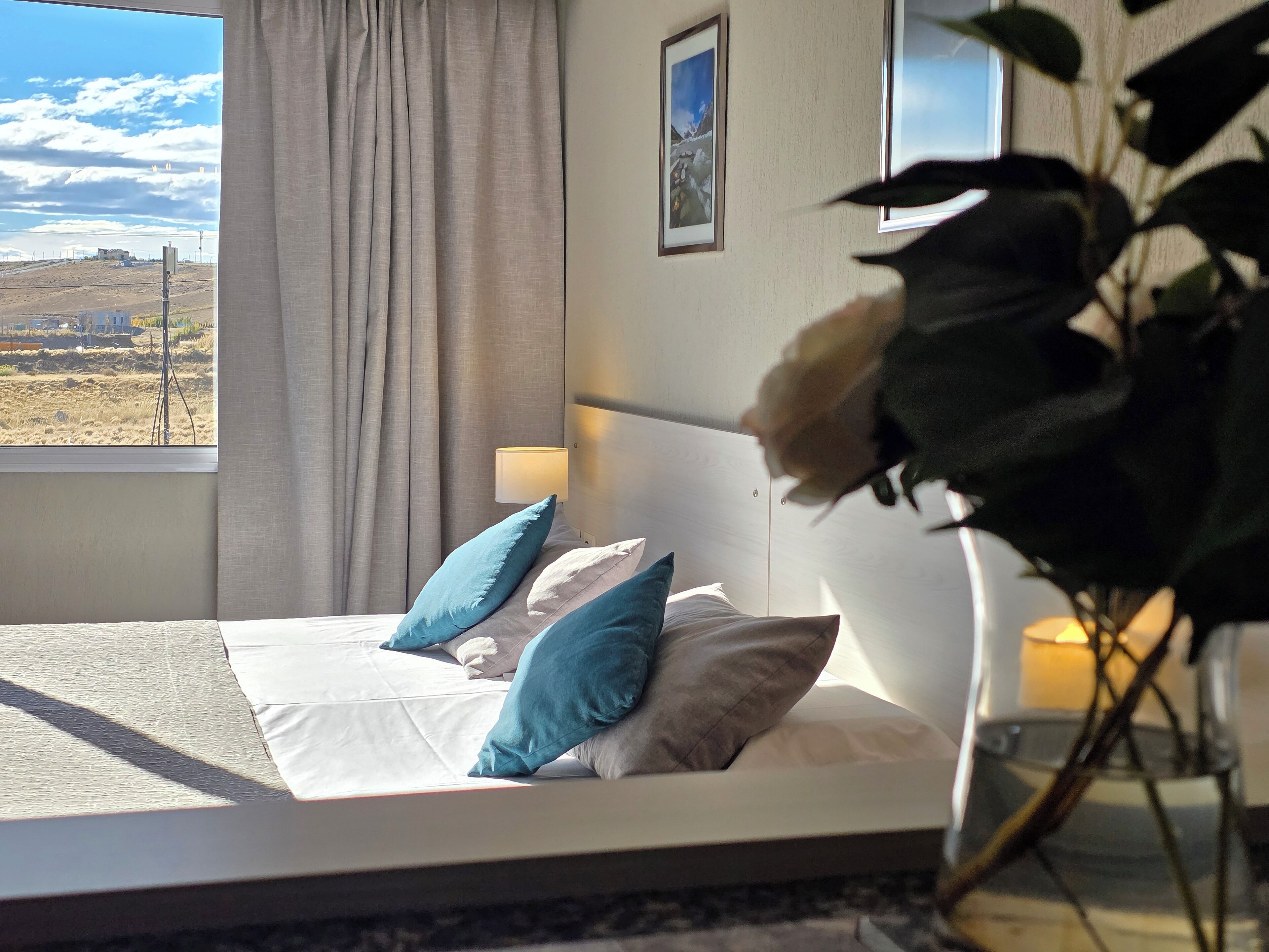 Un dormitorio con cama, almohadas, una mesita de noche, lámpara, ventana con vistas al paisaje y un cuadro en la pared.