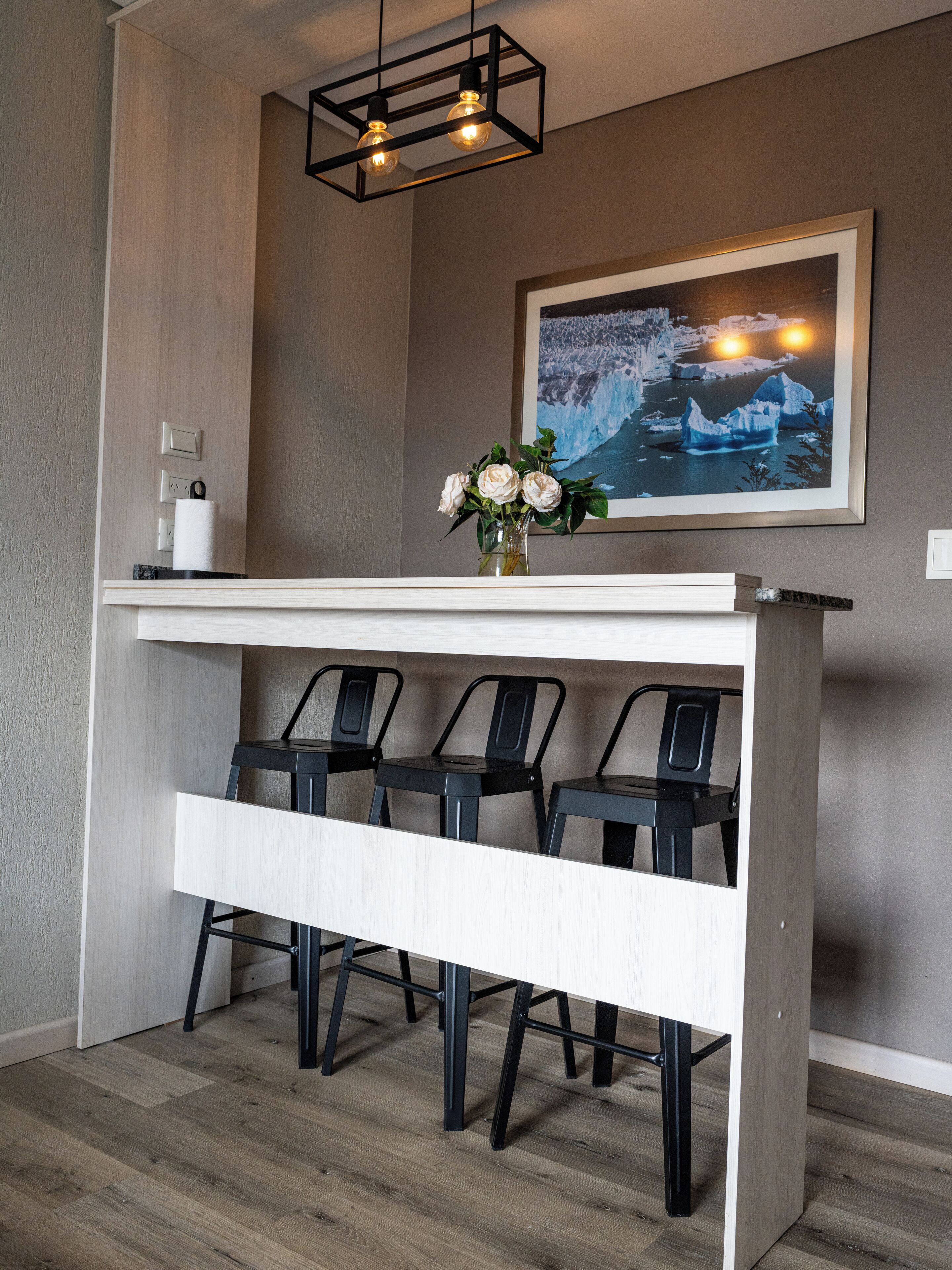 A modern kitchen with a white countertop, three black chairs, a vase with flowers, and a painting of icebergs on the wall.