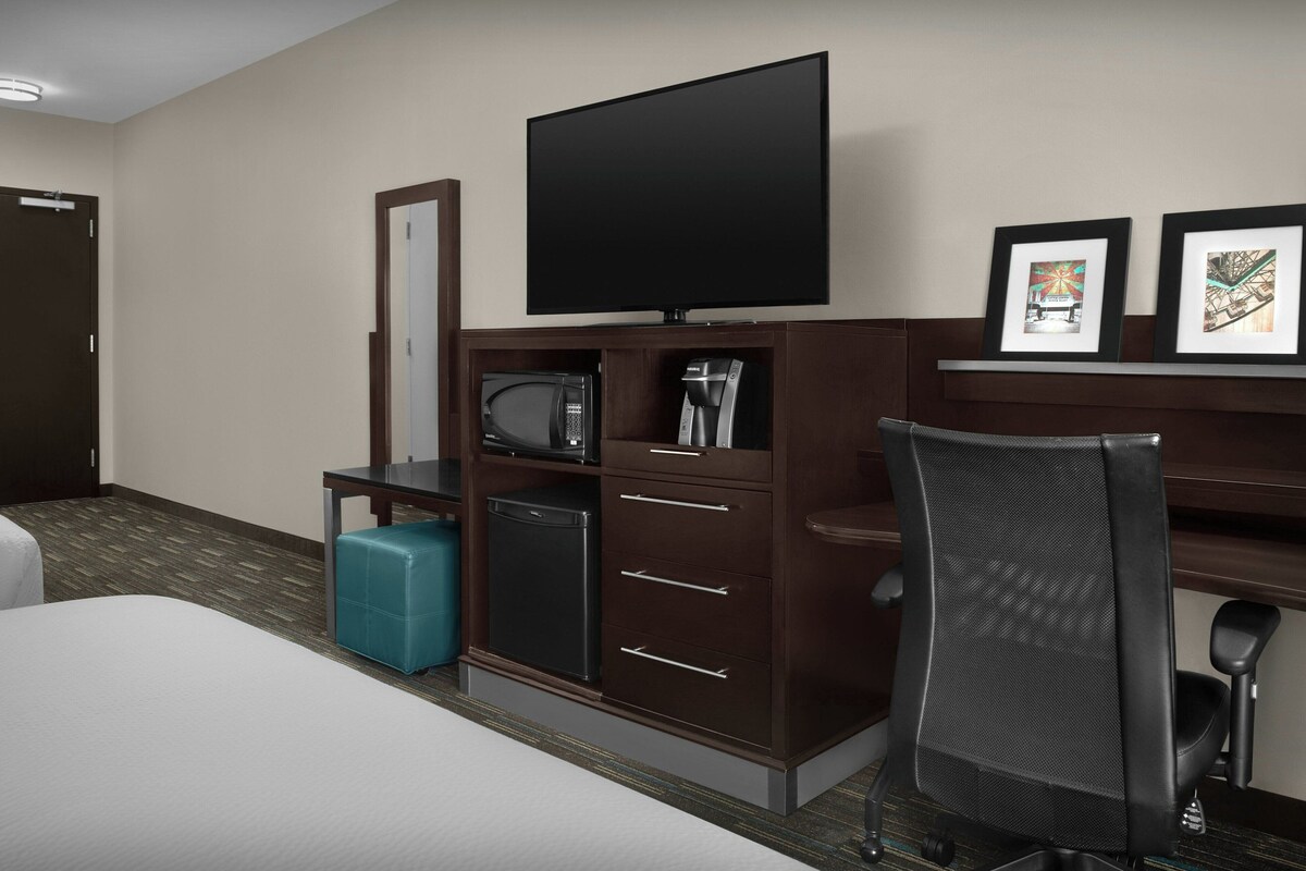 A hotel room with a bed, a desk with a chair, a television, a minibar, and a small trash bin.