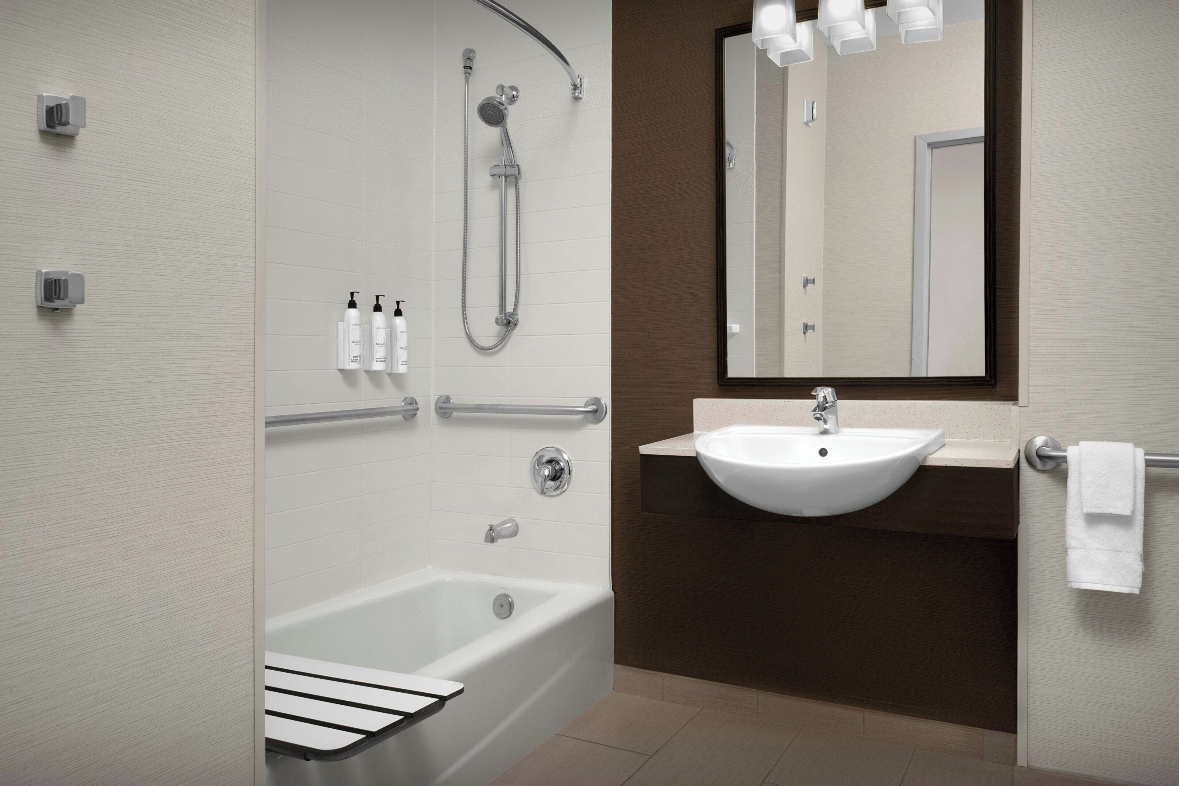 Une salle de bain moderne équipée d’une baignoire, d’une douche et d’un lavabo, ainsi que de barres d’appui et d’un miroir.