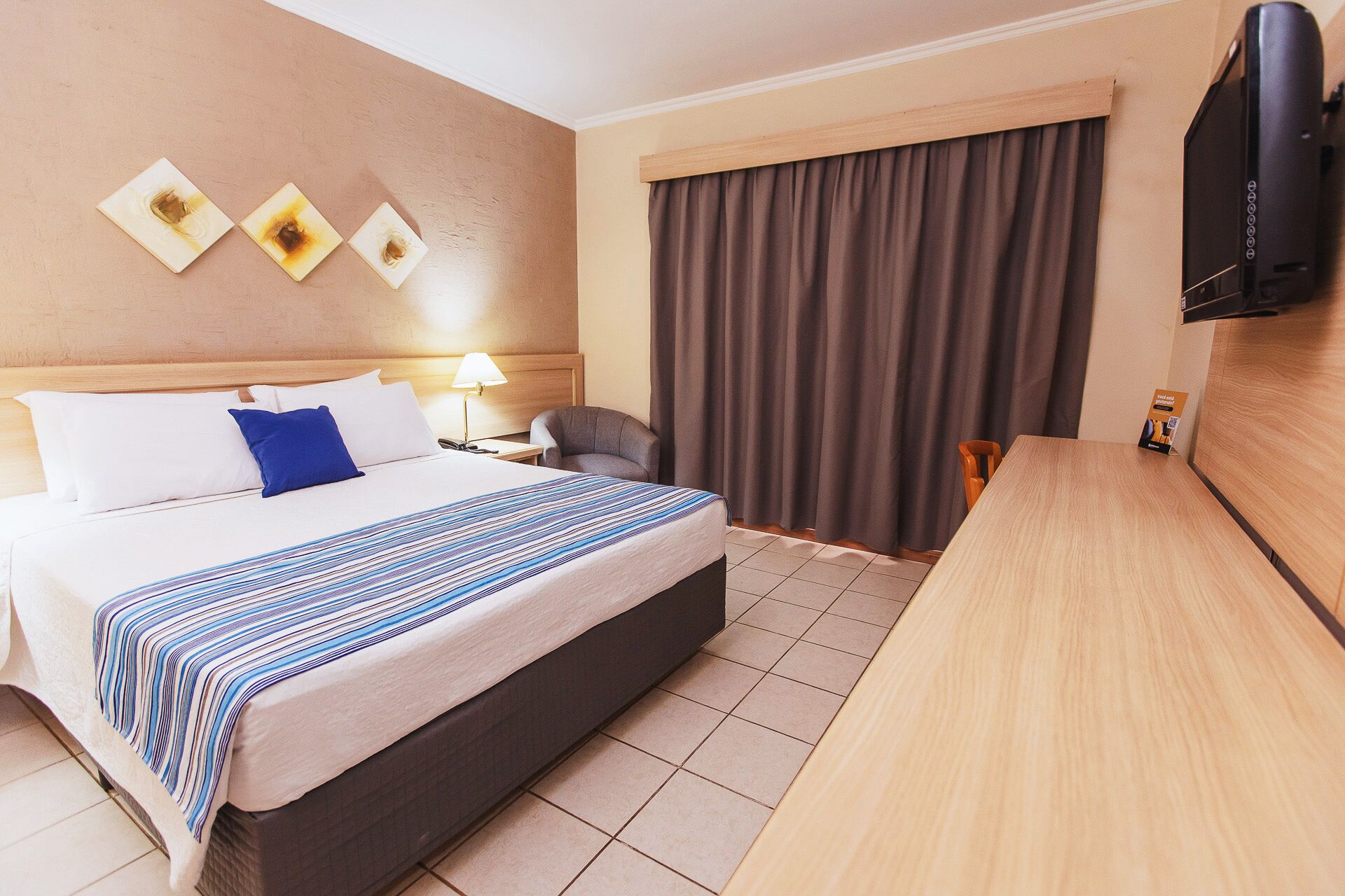 Habitació d'hotel amb llit, escriptori, cadira, televisió i finestra amb cortines.