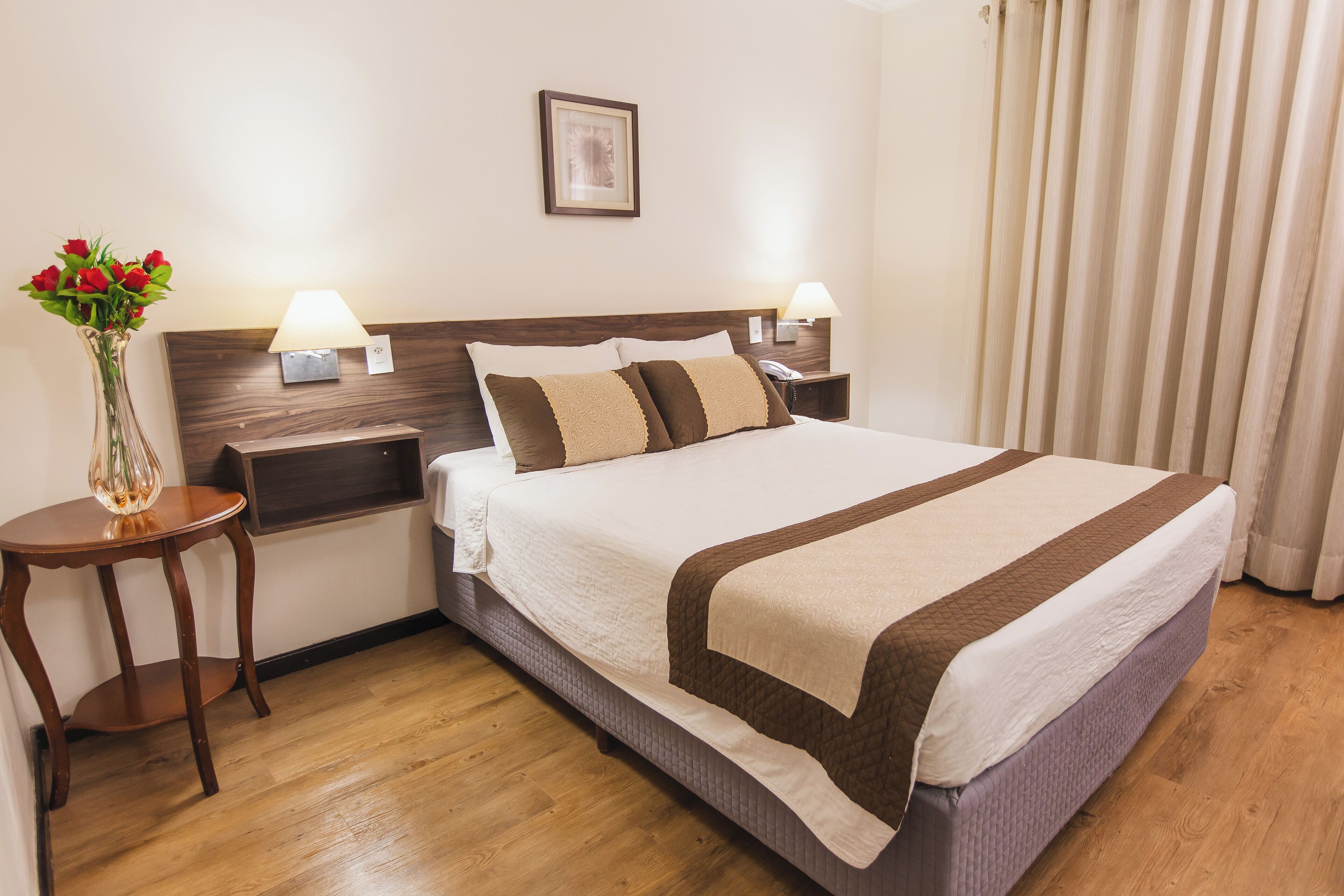 Une chambre d’hôtel bien rangée, avec un grand lit, deux tables de chevet dotées de lampes, un vase de fleurs et un tableau encadré au mur.
