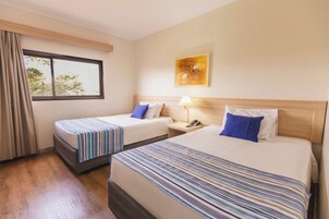 Habitación de hotel con dos camas, una ventana con cortinas, una mesita de noche con lámpara y un cuadro en la pared.