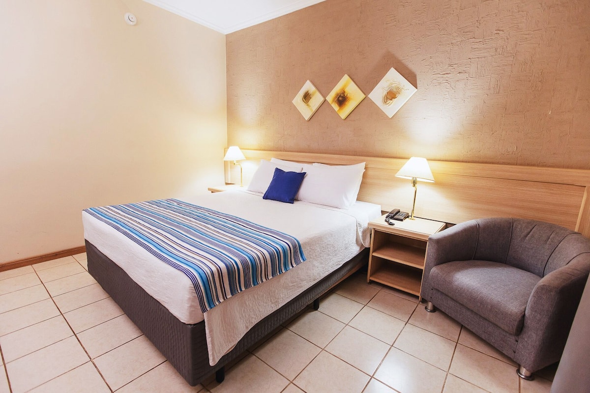 A hotel room with a bed, two bedside lamps, a nightstand, a chair, and three wall-mounted artworks.